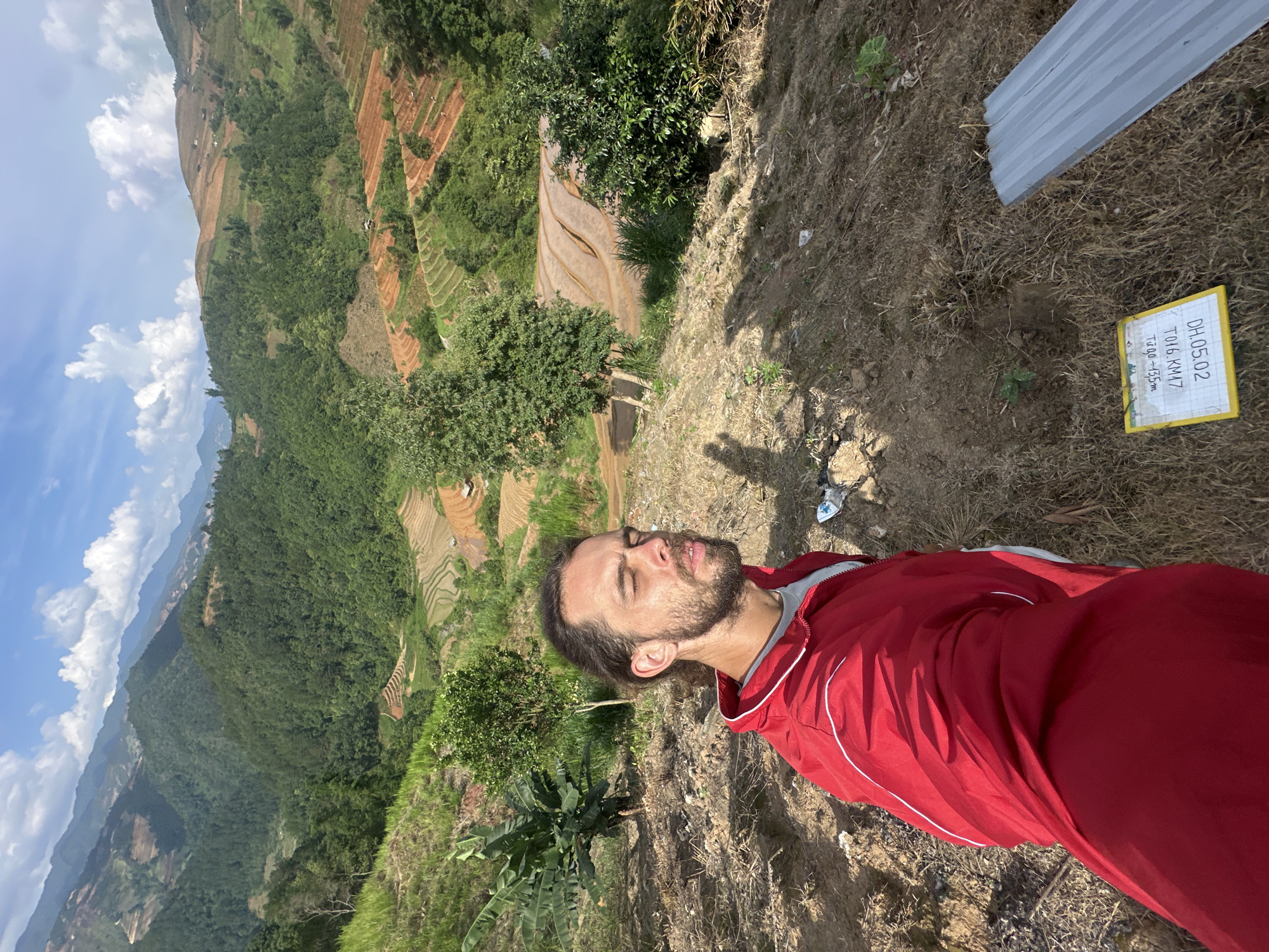 The image, oriented correctly, depicts a man resting in a mountainous landscape, likely in Lao Chai, Vietnam.
The main subject is a man with a short beard and dark hair, wearing a red long-sleeved athletic jacket over a grey t-shirt. He is positioned on a dry, uneven dirt and grassy slope in the foreground, with his eyes closed, head tilted slightly back, and face upturned towards the sky, suggesting relaxation or enjoyment of the sun.
The scene behind him is a vast and scenic vista. Rolling mountains covered in dense green foliage stretch into the distance under a bright blue sky with scattered white clouds. Carved into the hillsides are extensive terraced fields, some appearing to be recently tilled or flooded with water (displaying reddish-brown earth tones), while others are green with vegetation, possibly young rice plants. These terraces create intricate patterns across the landscape.
In the immediate foreground, next to the man, the ground is dry earth with patches of sparse grass and small plants. A grey corrugated metal sheet lies on the ground to the right. Also on the ground, slightly further right, is a yellow rectangular sign with a white border. The visible text on the sign reads:
DH.05.02
T016.KM17
Tự do + 15m
The overall setting is a high vantage point, offering panoramic views of the valleys and distant peaks. The day is bright and sunny, indicated by the clear sky and visible shadows, likely late morning or early afternoon. The terraced fields are a strong visual cue for the region. The man appears to be taking a peaceful break amidst the stunning natural beauty.