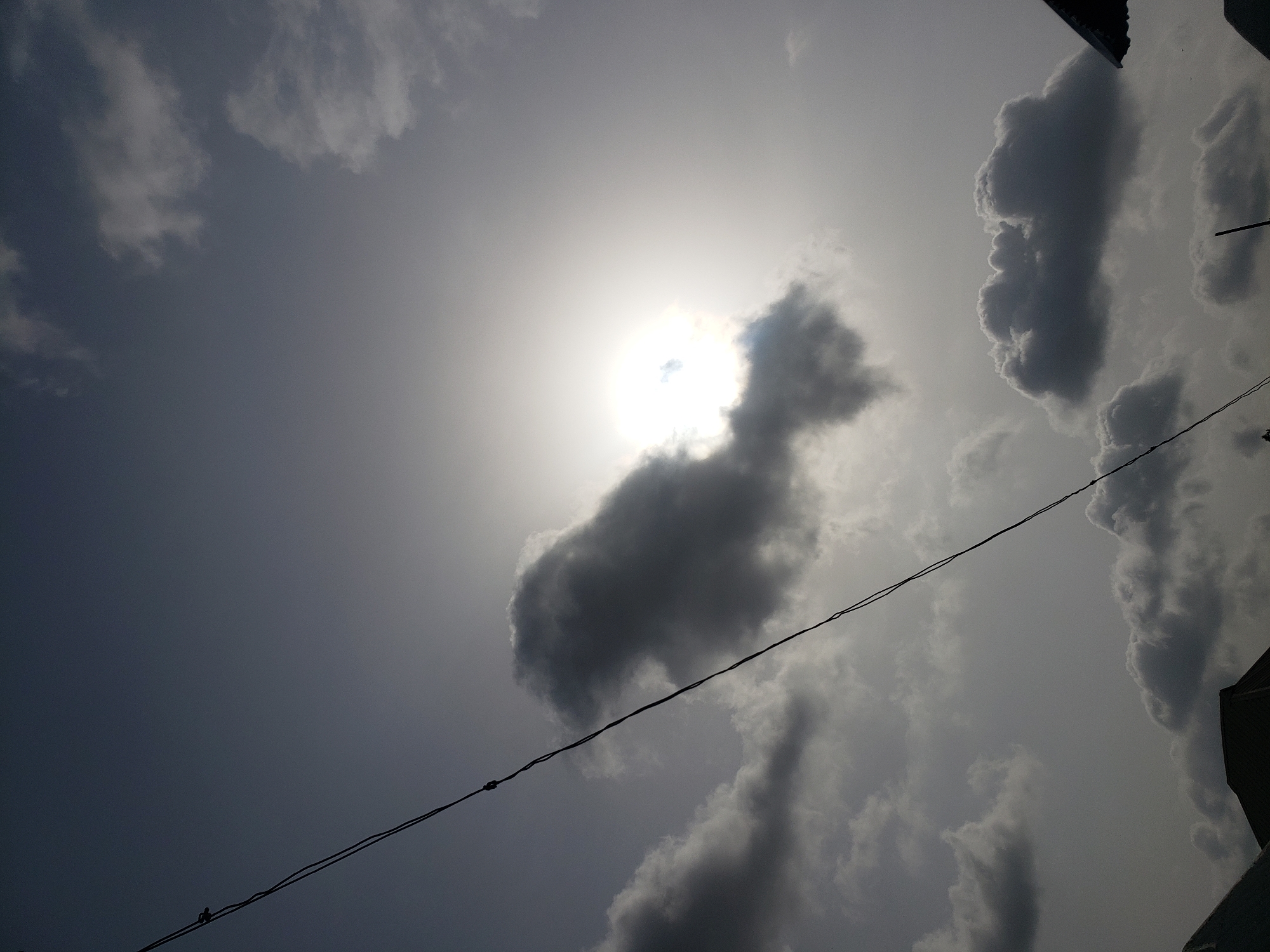 The image shows a daytime view of the sky in Adonkolo, Nigeria. The sun is visible through thin clouds, with darker cloud formations scattered throughout the frame. A power line runs diagonally across the lower portion of the image, and the corner of a building is visible in the lower right corner. No people are visible.