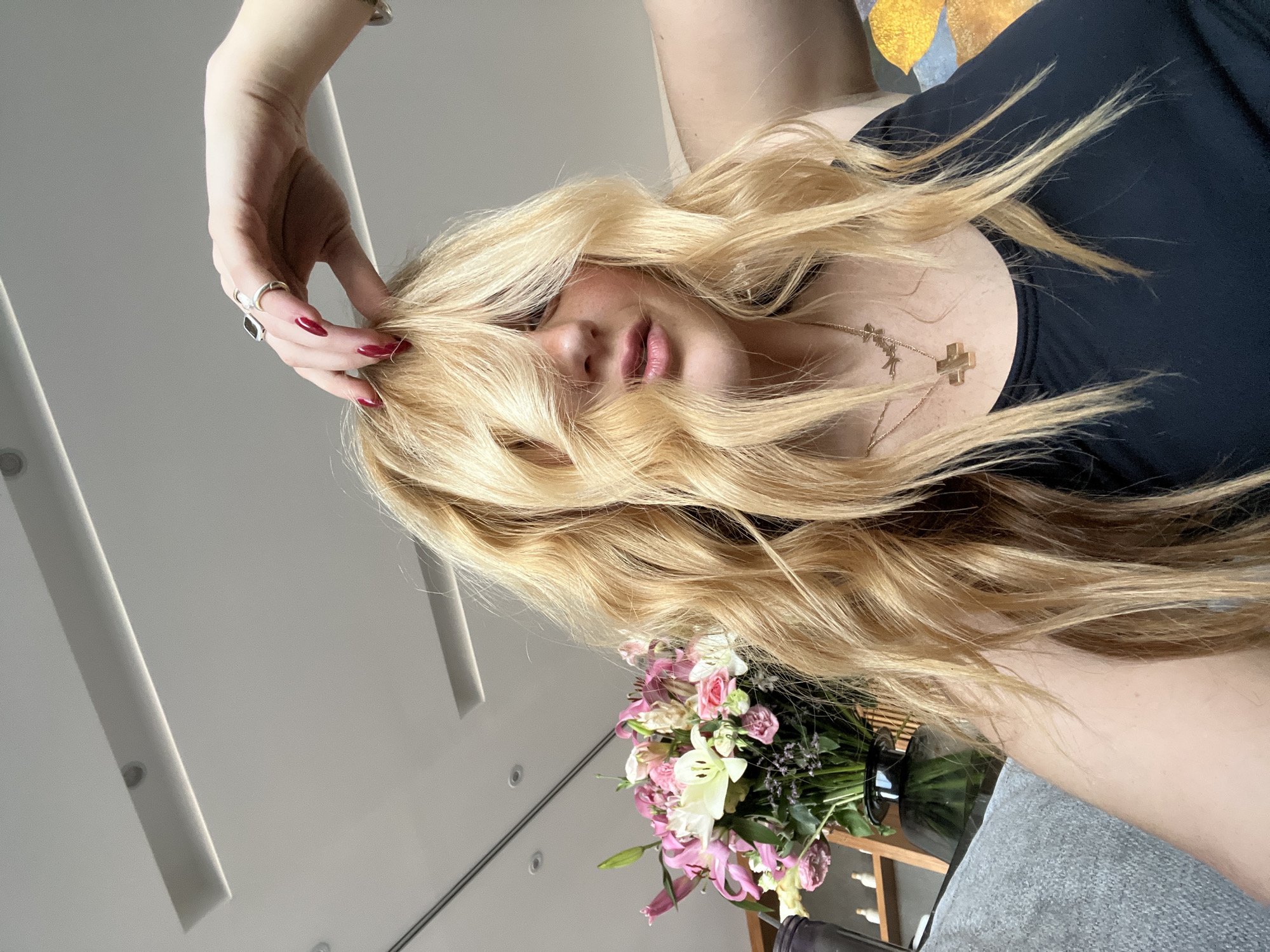 An indoor, close-up photograph captures a blonde-haired individual, presumed female, with wavy hair partially obscuring her face. Her left hand, featuring dark red manicured nails, is raised to her forehead, fingers touching her hair. She wears two rings on her left hand: one with a dark, rectangular stone on the middle finger and a simpler band on the ring finger. A silver-toned bracelet is visible on her left wrist. The individual is clad in a black tank top, displaying two gold-toned necklaces: one with a cross pendant and another with small, abstract bird-like charms. In the background, to the left of the subject, a bouquet of pink and white lilies, roses, and other flowers is arranged in a dark vase on a surface. Further left, a wooden shelving unit holds several small, light-colored decorative objects. To the right, a textured gray upholstered surface is visible, alongside a blurred piece of art with yellow and blue hues. The ceiling features integrated rectangular and circular light fixtures. The scene is located within a modern interior in Santiago Yancuitlalpan, Mexico.