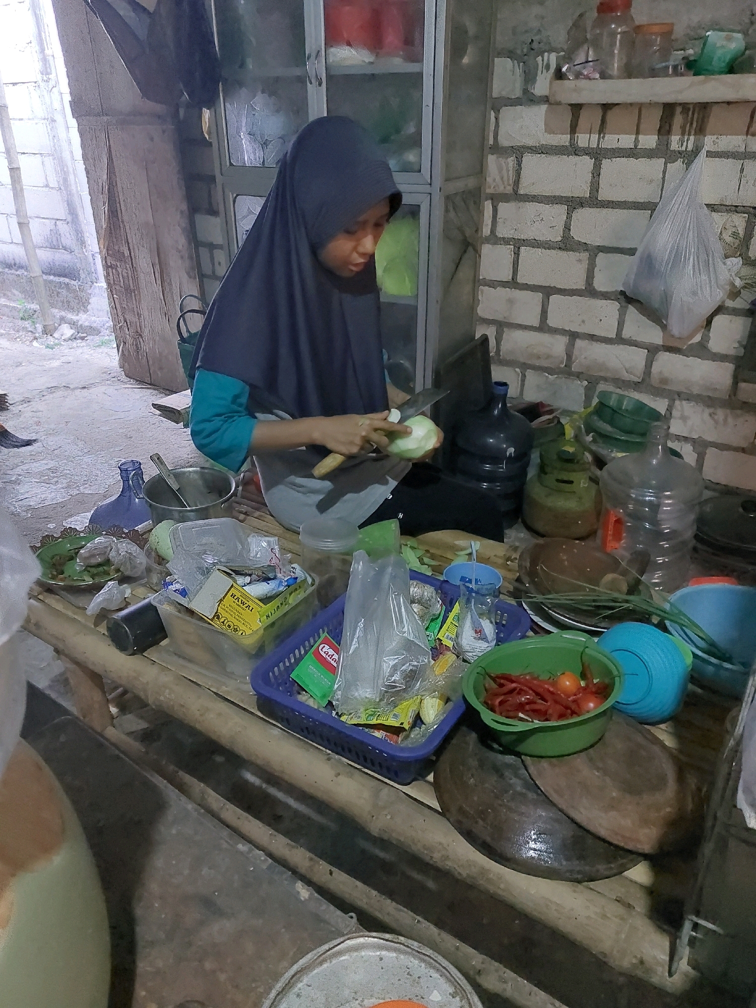 A person wearing a dark blue hijab is chopping vegetables in a rustic kitchen in Sarangan, Indonesia. The kitchen appears to be outdoors or in an open-air structure, with a bamboo table serving as a workspace. Various ingredients and cooking utensils are laid out on the table, including chilies, tomatoes, plastic bags of food, a metal pot, and a knife. In the background, brick walls and large water jugs are visible. The scene suggests a simple, communal cooking environment.
