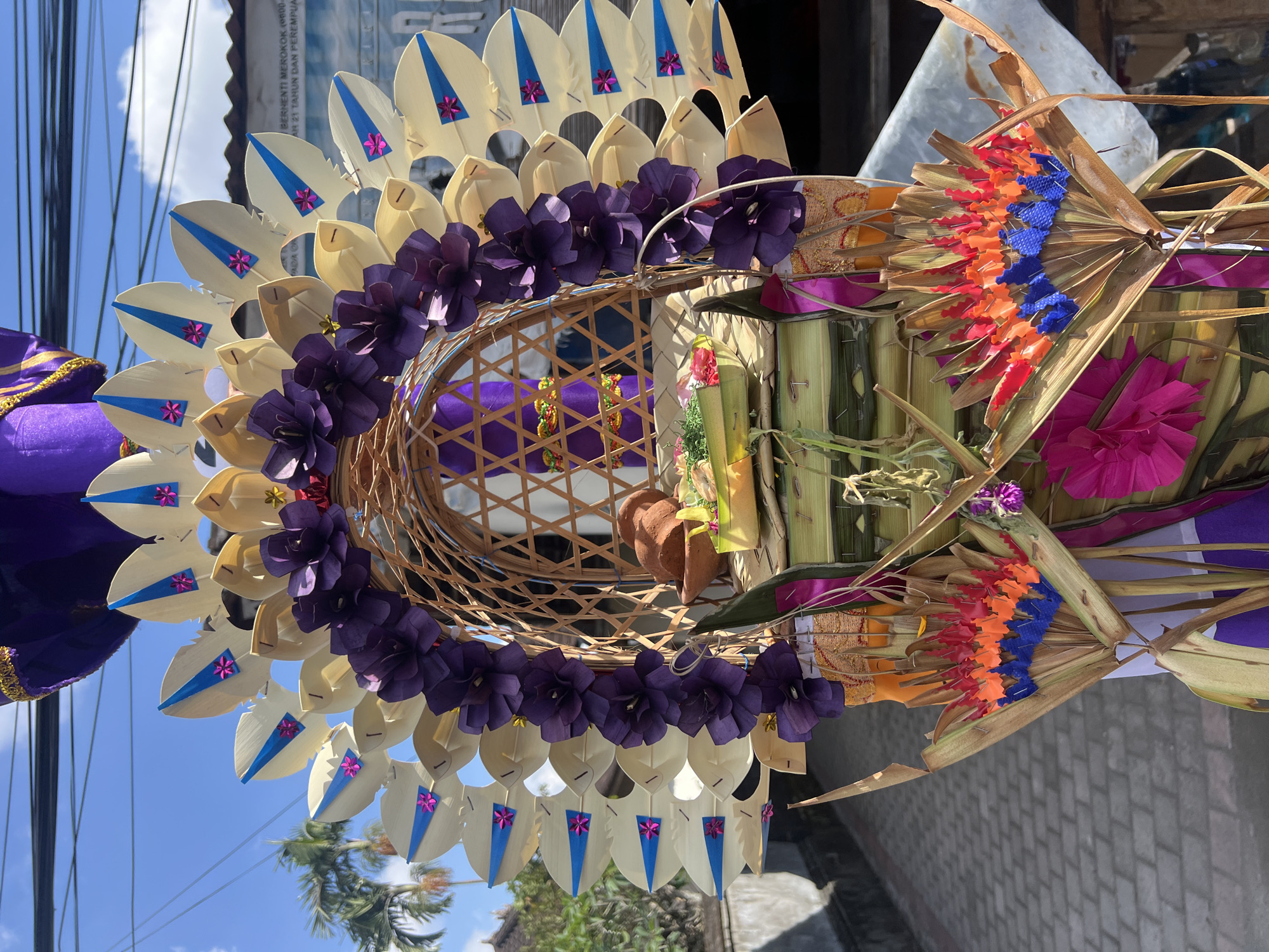 This is a medium shot, eye-level, outdoor photograph of a colorful Balinese offering, likely a "gebogan" or "canang sari" arrangement, placed on a paved street. The offering is elaborately decorated with various natural materials and artificial embellishments. Dominating the center is a woven bamboo basket, housing smaller offerings of food items and a clay pot. The basket is framed by large, petal-like structures made of dried palm leaves, accented with blue and pink details. Purple paper flowers are draped around the top of the basket, and further down, there are fan-shaped arrangements made of dried leaves with colorful, fringed inserts in red, orange, and blue. The offering is rich in texture and color, with layers of green bamboo leaves and fabric, including purple, orange, and pink elements, forming its base. In the background, a building with a tiled roof and an overcast sky are visible. Palm trees are also present in the distance. The scene appears to be set in a residential or street area within the city of North Kuta, Indonesia, given the architectural style and vegetation. There are no people visible in the photograph.