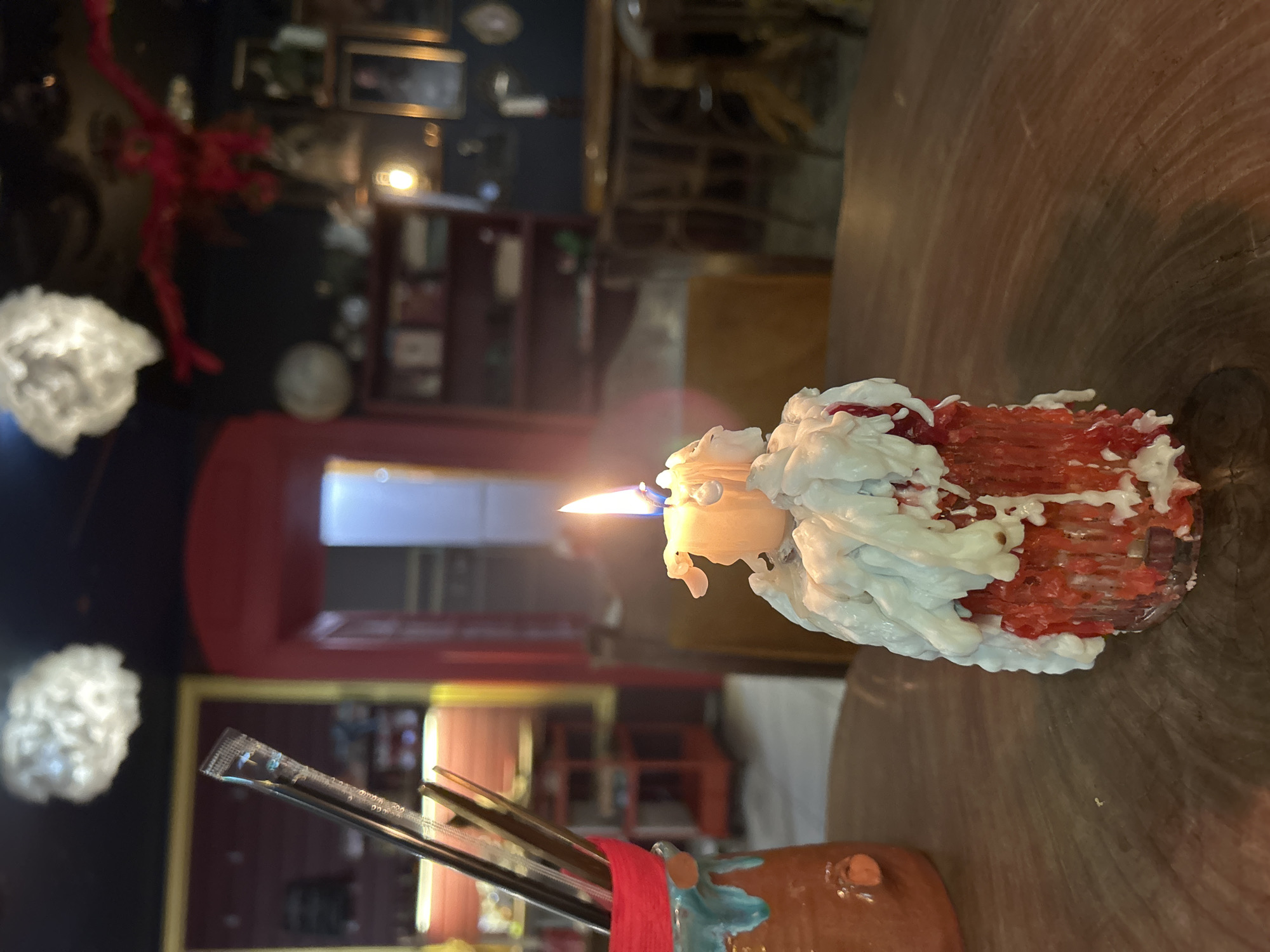 A lit candle, with significant wax drippings, sits on a wooden table. The candle is red at the bottom and cream-colored wax drips down its sides, some of which are tinged red. Behind the candle, a blurred background shows a room with a red archway, bookshelves, and chairs, suggesting an indoor setting such as a restaurant or bar. To the left of the candle, a ceramic holder with a red rim contains several straws, one of which is partially in its packaging. The overall lighting is dim, with the candle flame and a few overhead lights providing illumination.