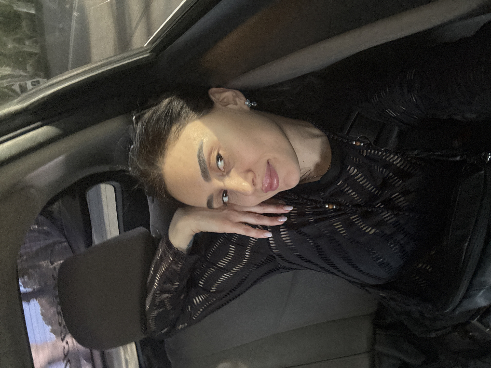 A woman is resting her head on her hand in what appears to be the back seat of a car. The background, visible through the car window, suggests an urban environment, potentially at night, and likely in Tbilisi, Georgia. The woman is wearing a black, possibly sheer, top. The car interior is dark, with a headrest visible to the left of the woman.