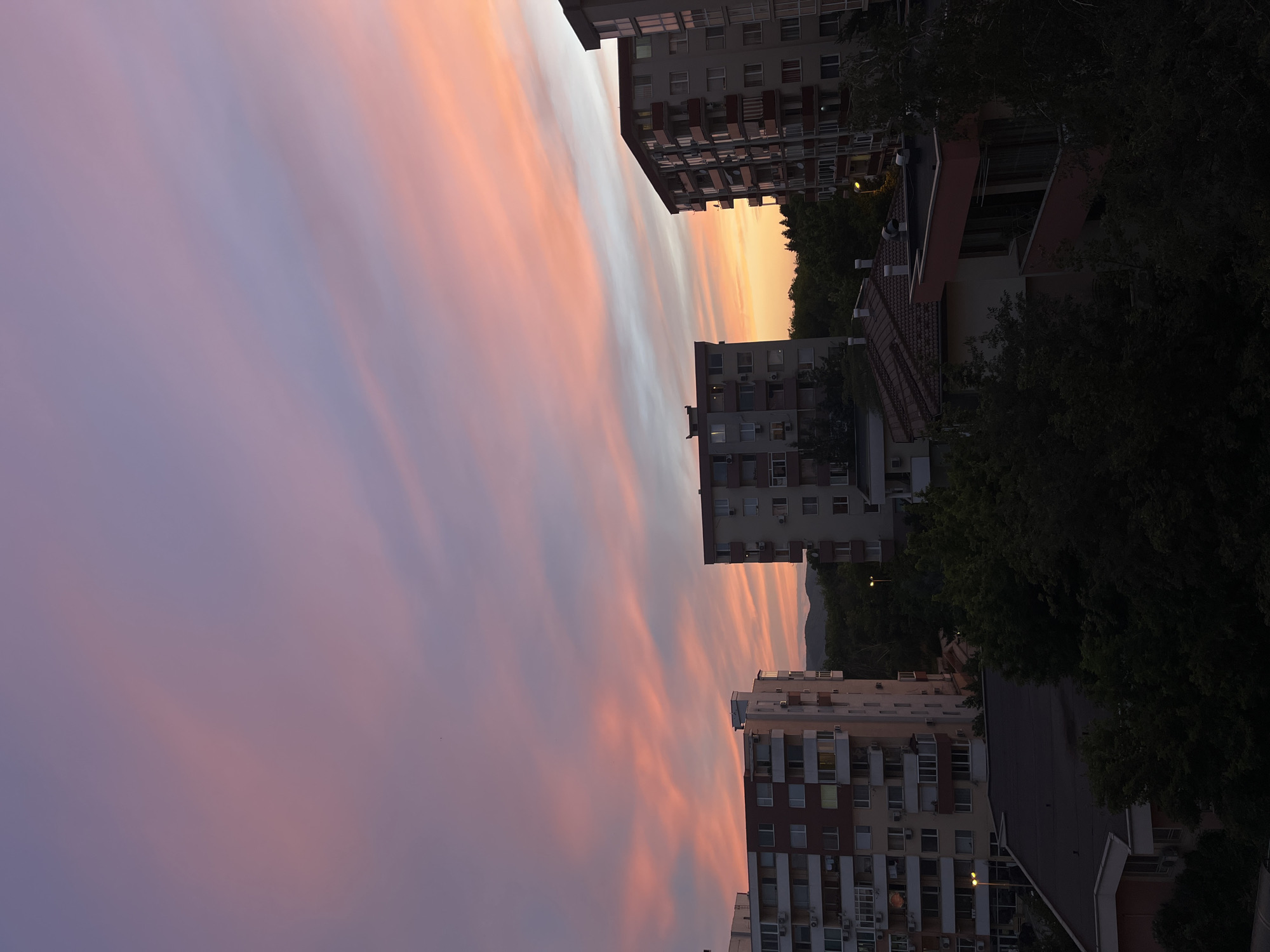 This is a photograph of a city skyline at sunset in Veliko Tarnovo, Bulgaria. The sky is a vibrant mix of pink, orange, and purple hues, with wispy clouds stretching across the expanse. In the foreground, several apartment buildings are visible, characterized by their repetitive window patterns and balconies. The buildings vary in height and architectural style, with some featuring darker colored facades. Lush green trees are interspersed between the buildings and in the foreground, indicating a well-vegetated urban area. In the distance, faint outlines of hills or mountains can be seen against the colorful sky. The lighting suggests it is either early morning or late evening, with artificial lights from some windows and street lamps beginning to illuminate. No people or specific actions are discernible in the image.