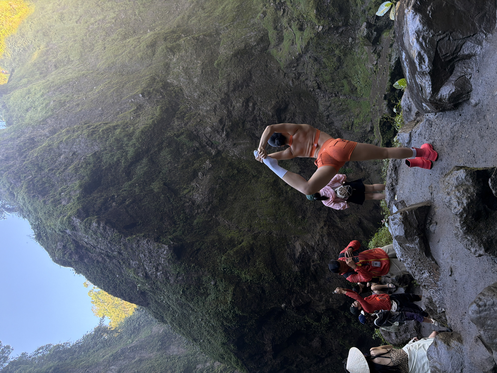 In Sidomulyo, Indonesia, a woman performs a yoga pose resembling a dancer's pose, balancing on one leg with the other foot held behind her head. She wears an orange sports bra and matching shorts, white socks, and bright pink boots. She stands on a muddy, rocky path within a narrow canyon where steep, moss-covered walls rise dramatically on either side, forming a natural enclosure. Sunlight filters down from above, illuminating parts of the canyon and creating a bright haze. Several other individuals are visible in the background, some appearing to be tourists, one of whom is taking a photo with a phone. The environment is characterized by rugged terrain, lush vegetation clinging to the rock faces, and a damp, natural atmosphere.