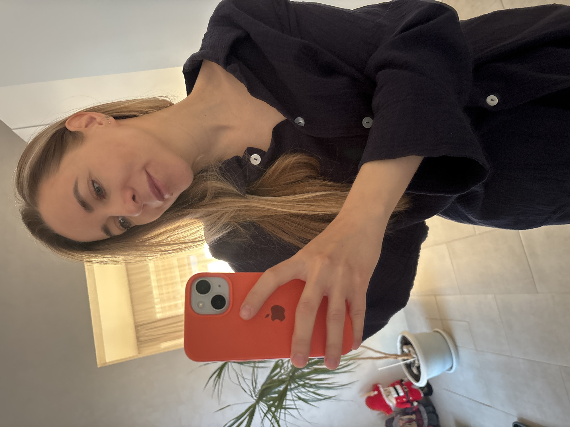 A mirror selfie captures a young woman with long blonde hair and light-colored eyes, looking directly into the camera with a slight smile. She is wearing a dark blue or black textured long-sleeved shirt or cardigan, fastened with light-colored buttons. Multiple small stud earrings adorn her left ear. She holds an orange iPhone with a red case, prominently displaying the Apple logo, in her right hand, angled towards her reflection.
The setting appears to be an indoor space, likely a home, characterized by a light-colored, possibly off-white, tiled floor. A light-colored wall serves as the background, and a window with horizontal blinds is reflected in the upper right portion of the image. On the floor to the right, a potted green plant with thin leaves is visible, along with a partially obscured red and white figurine, resembling a Santa Claus or similar holiday decoration. The lighting suggests it is daytime. The location context for this media is Chaikovsky, Russia.