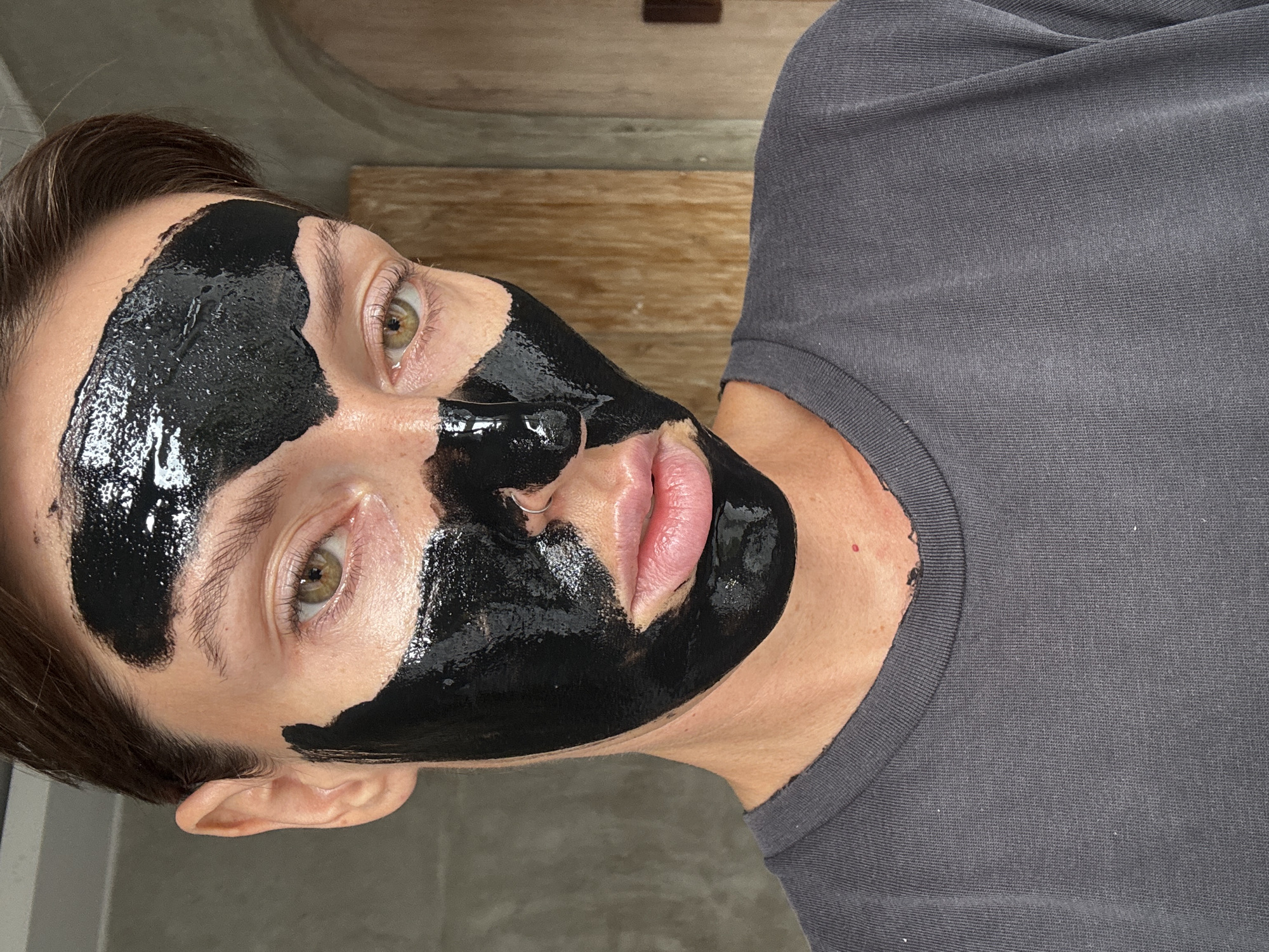 A close-up shot shows a person with a black face mask on their face. The mask covers most of their forehead, cheeks, and nose, with portions of their eyes and lips visible. The person is wearing a gray t-shirt. The background is out of focus, but appears to be an indoor setting with wooden paneling. This image was taken in North Kuta, Indonesia.