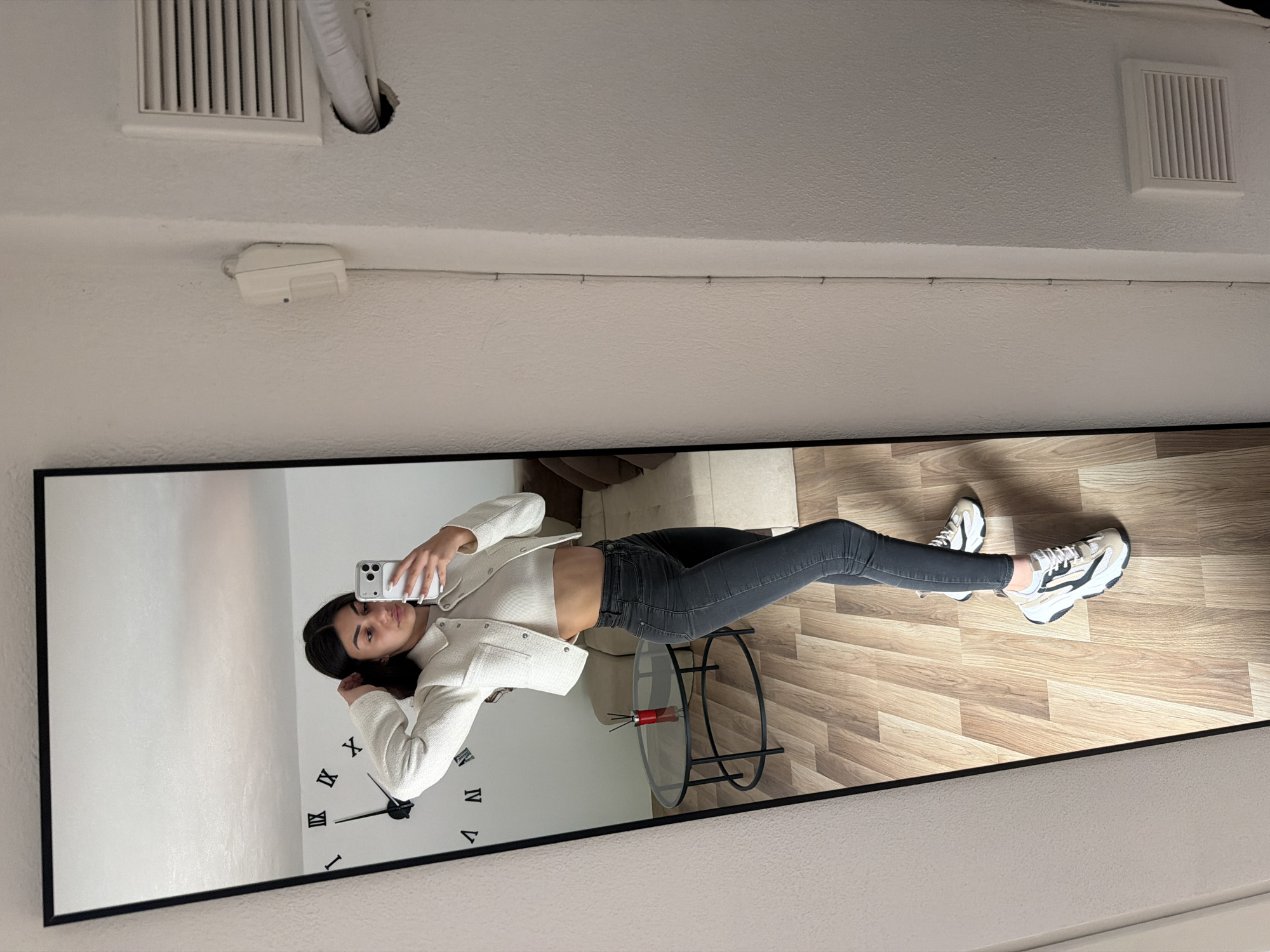 A person is standing in front of a full-length mirror, taking a selfie with their phone. The mirror's reflection shows the individual wearing a white jacket, a white top, gray jeans, and white sneakers. The mirror also reflects a clock on the wall behind the person, a glass table with a red object, and a wooden floor. The wall in the background is white. The location is within a residential dwelling. There is no evidence to suggest this location is in Gabrovo, Bulgaria. Additional elements in the room include ceiling vents and a security sensor.