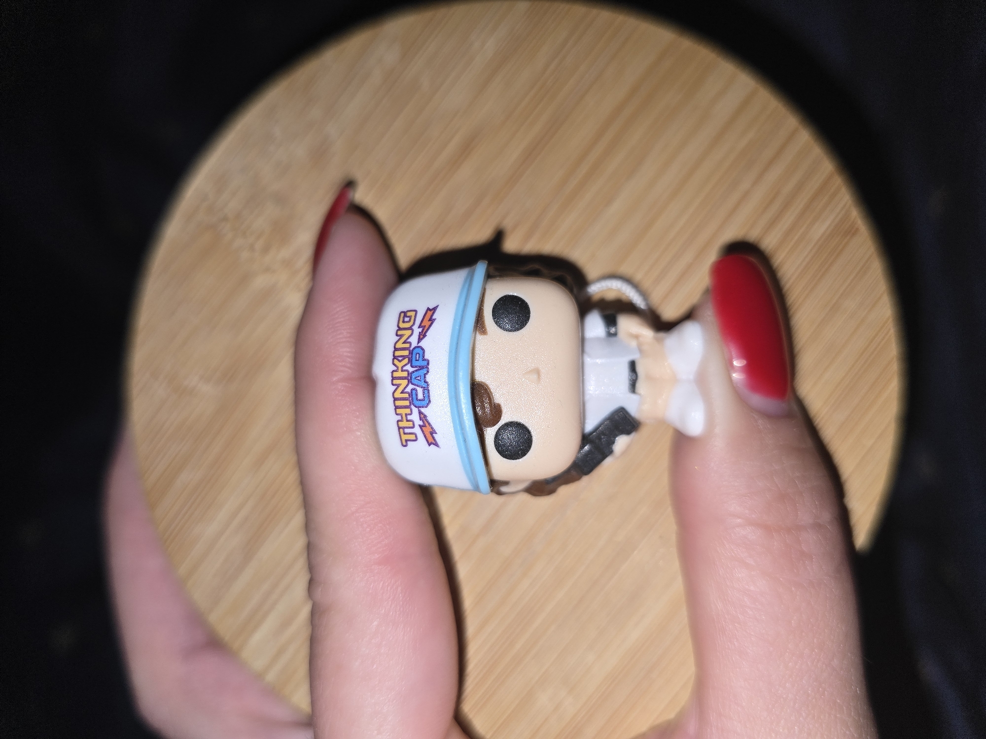A close-up shot shows a person's fingers, with red nail polish, holding a small toy figurine on a round, light-brown wooden surface. The figurine is a miniature character wearing a white cap with blue trim and the words "THINKING CAP" in colorful letters. The figurine has black eyes, a pale face, and is dressed in a white outfit. The wooden surface appears to be the lid of a container and has visible wood grain. The background is dark, suggesting low lighting conditions. The location is not specified in the image or the provided context.