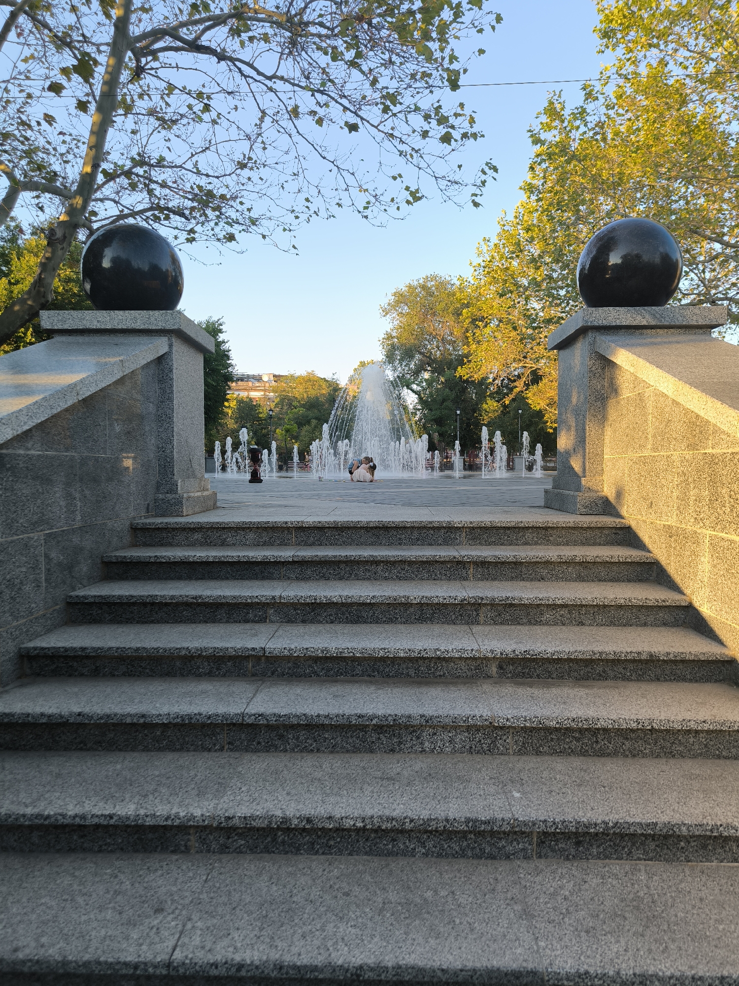 This image depicts an outdoor scene in Mykolaiv, Ukraine, featuring a public square with a fountain and surrounding trees. In the foreground, a wide granite staircase leads up to the square. On either side of the top of the stairs, there are stone pillars topped with large, dark, spherical sculptures. The square itself has a paved surface, and a fountain with multiple jets of water is active in the mid-ground. Two individuals, appearing to be a man and a woman, are seated near the fountain, with the woman facing away from the camera and the man partially visible. In the background, trees with yellowing leaves suggest an autumn setting, and buildings are visible beyond the trees. The lighting indicates it is late afternoon or early evening, with the sun casting long shadows.
