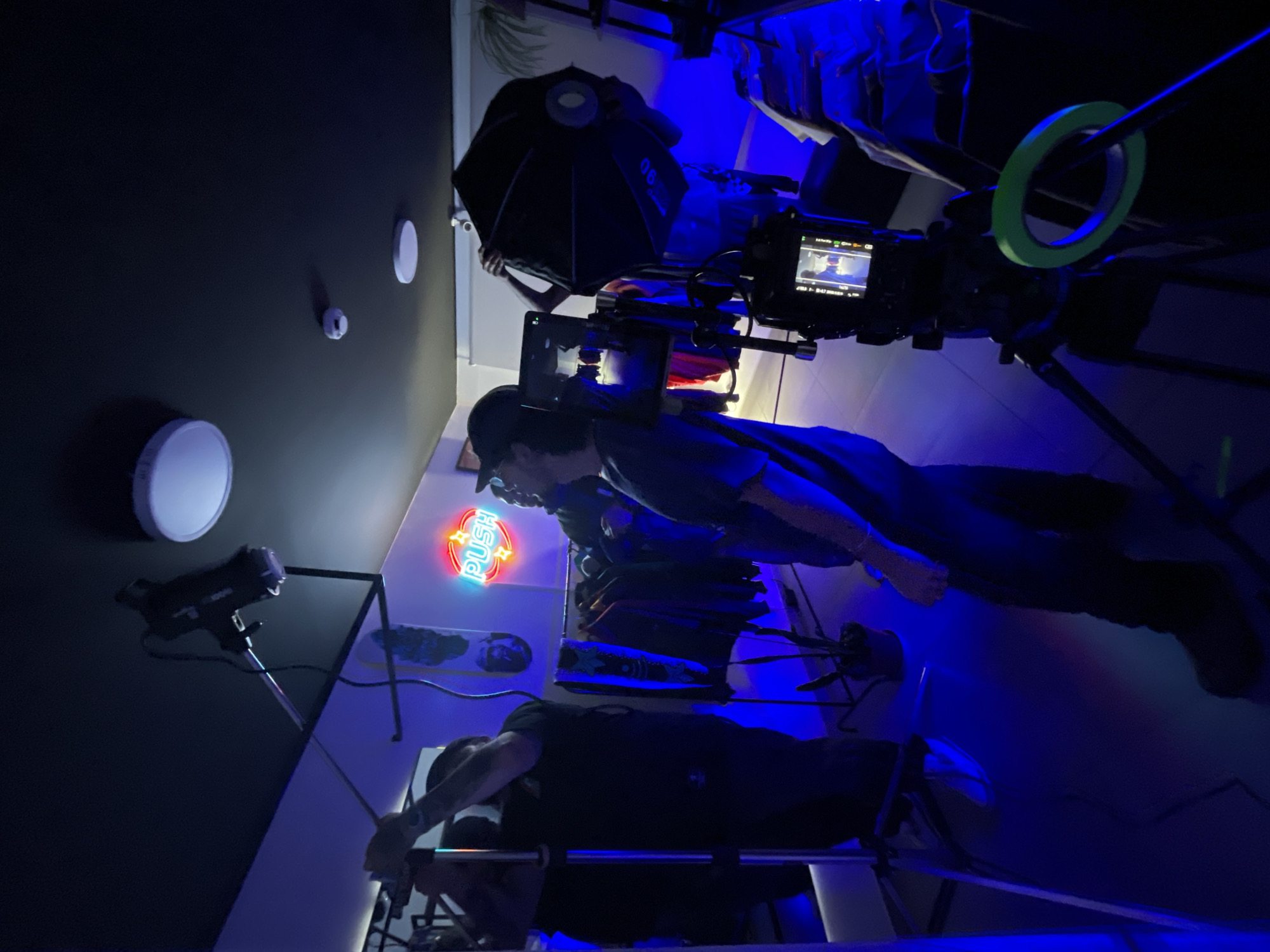 The scene appears to be a filming setup in an interior space with blue lighting. There are two individuals visible, one in the foreground performing an action on what looks like exercise equipment, and another partially visible behind a camera setup. The camera rig includes a screen, a circular green object (possibly tape), and an umbrella reflector. Behind the individuals and equipment, there is a neon sign reading "PUSH", skateboards mounted on a wall, and several ceiling-mounted light fixtures. The location is not discernible from the image.