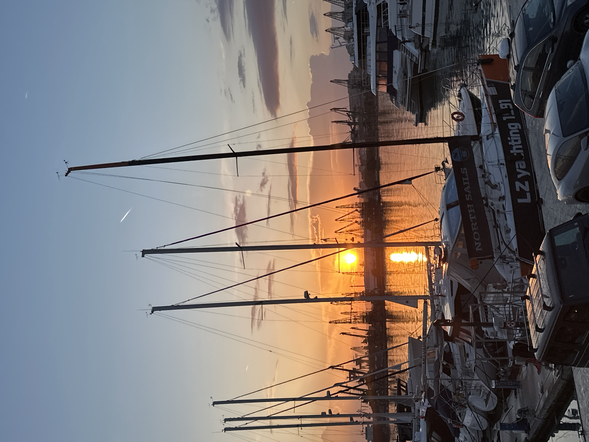 The image shows a marina in Varna, Bulgaria, at sunset. Multiple sailboats are docked in the foreground, with their masts rising into the sky. The setting sun reflects on the water between the boats, creating a bright orange glow. In the background, cranes are visible on the harbor, silhouetted against the colorful sky. Cars are parked along the edge of the marina next to some of the boats. Some of the sailboats have visible branding, including "NORTH SAILS" and "LZ yachting 1.91". There are thin clouds in the sky, tinged with orange and pink. The light is warm and diffused.