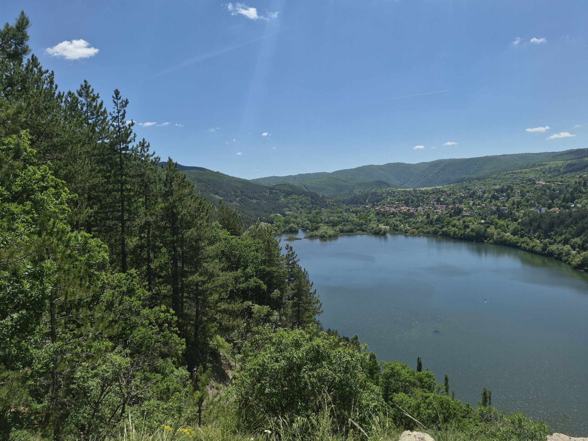 The image captures a serene, natural landscape featuring a sizable body of water, likely a lake or wide river, flanked by dense, verdant forests and gently rolling hills in the background. The sky is a bright blue, punctuated by a few scattered clouds. A village or small town is nestled amongst the greenery in the distance. The foreground is dominated by lush trees and vegetation, suggesting a vantage point from a hillside or elevated terrain. The scene evokes a sense of tranquility and natural beauty. Location details are unavailable.