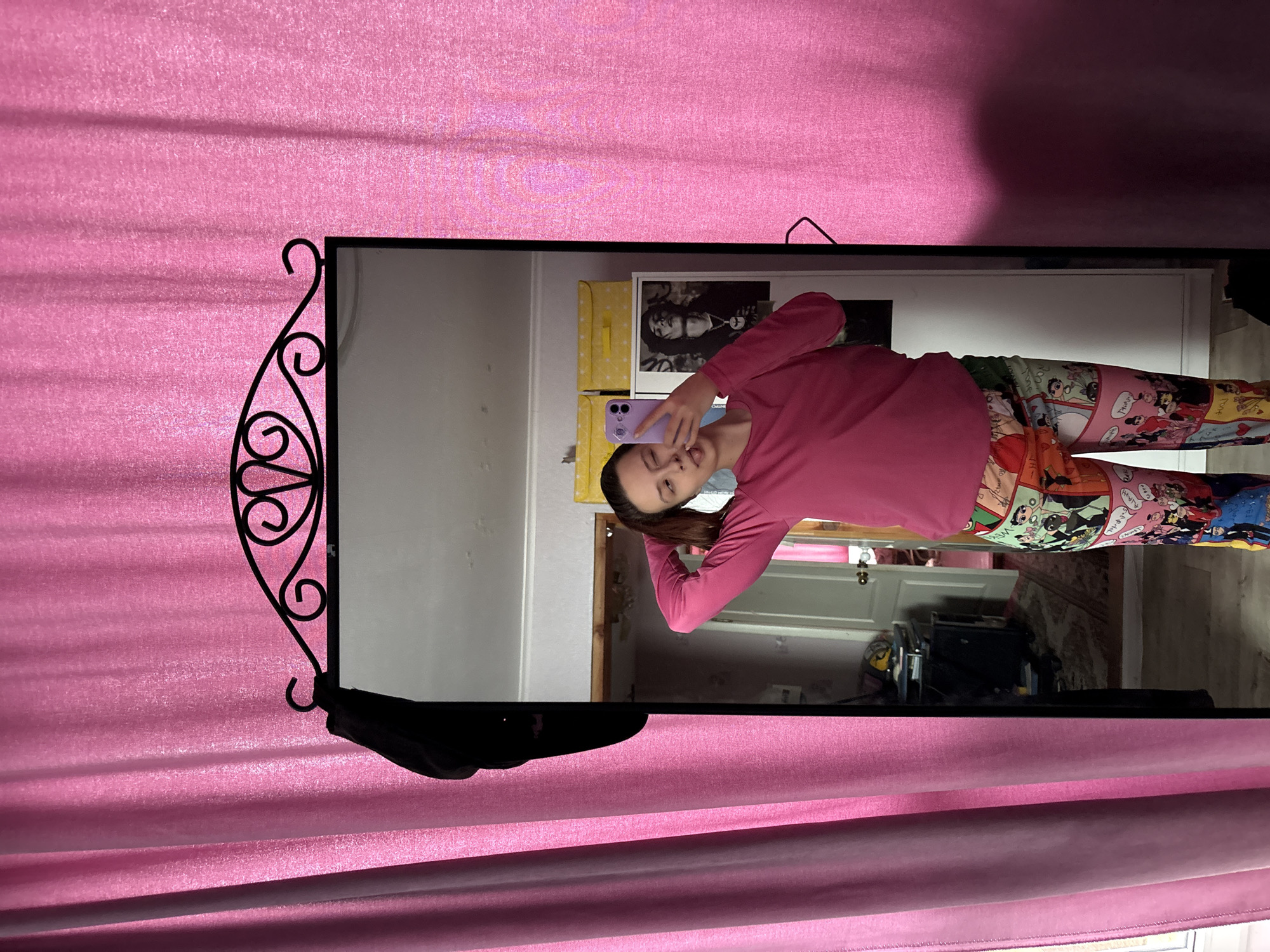 A young woman is reflected in a decorative mirror hanging in front of pink curtains. She is wearing a long-sleeved pink shirt and colorful leggings with cartoon images. She is holding a purple cell phone in her left hand and appears to be taking a selfie. Behind her in the reflection, there are cabinets, boxes, and a framed picture on a wall. A doorway leads to another room with furniture and a patterned rug. The overall location is unknown, and there is no evidence to indicate it's in the city of Прикамский, Russia.