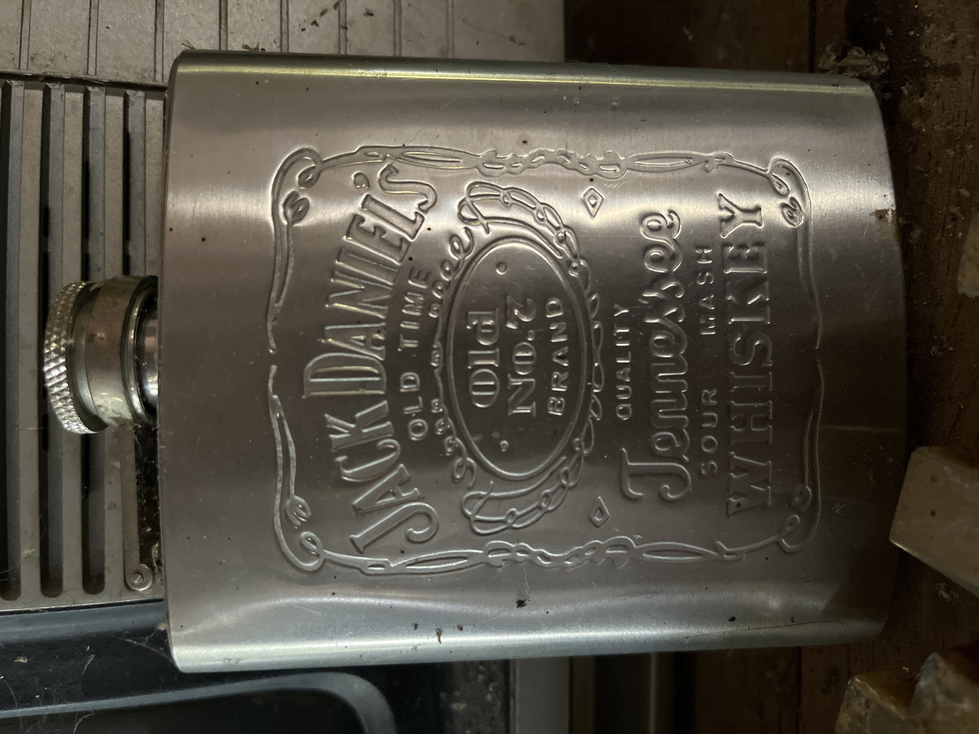 A close-up, eye-level, medium shot captures a silver-toned flask with embossed text. The flask is positioned vertically, with the cap at the top. The embossed text reads "Jack Daniel's," "Old Time," "Old No. 7 Brand," "Tennessee," "Sour Mash," and "Whiskey." The flask appears to be made of stainless steel and has a slightly worn appearance. In the background, horizontal grates are visible, suggesting the flask is resting on or near some kind of ventilation system. The bottom of the image shows a wooden surface with several small, yellowish-brown rectangular objects, possibly candies or small pieces of wood, scattered on it. The overall lighting is dim, with the flask reflecting the light and creating highlights. The scene is devoid of any individuals or discernible human activity. The flask's design and branding strongly suggest it is a container for alcoholic beverages. The image does not provide any specific geographical markers to pinpoint its location in Tambakreja, Indonesia.