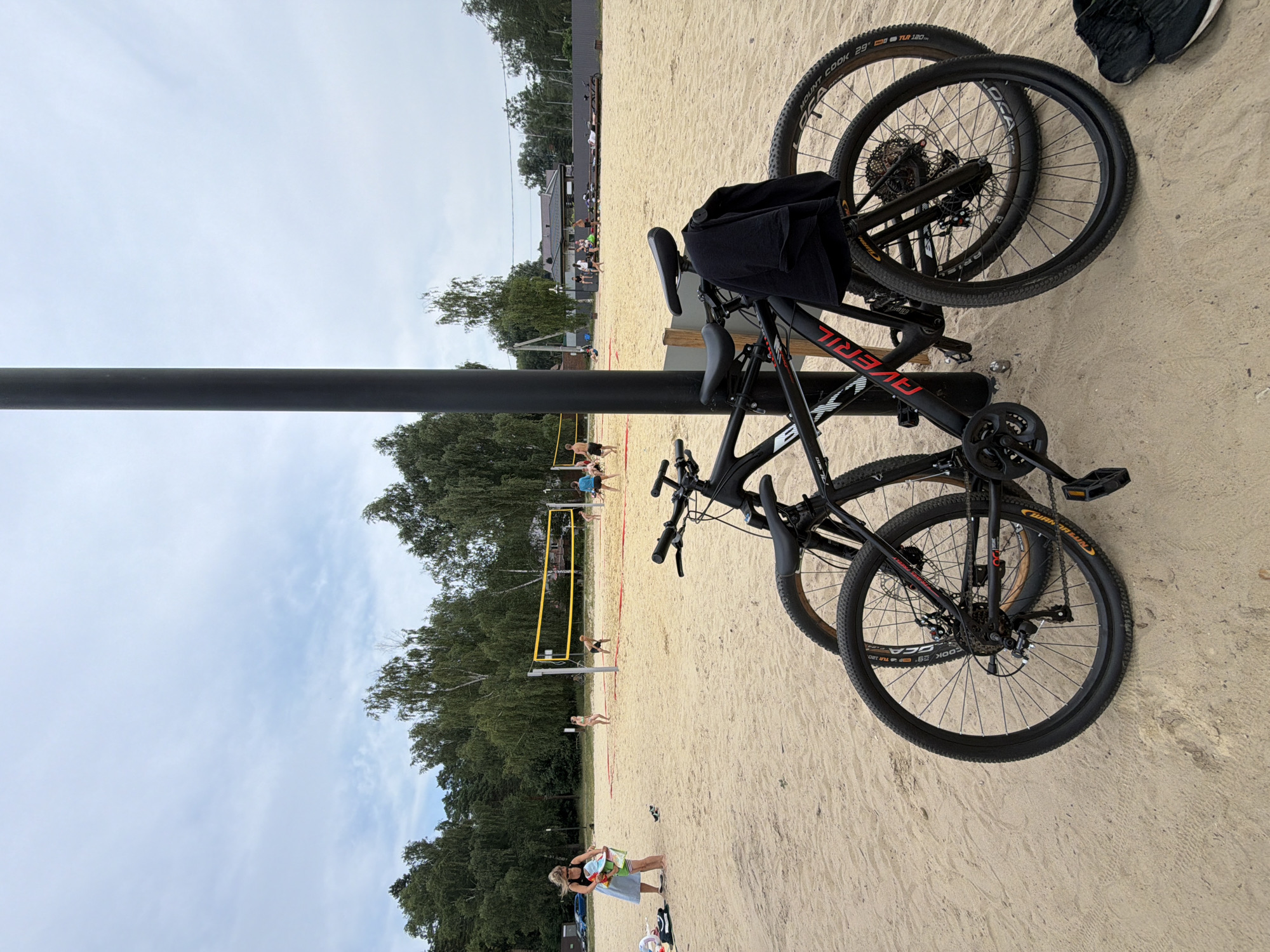 This is a medium shot of a beach volleyball court in Malakhovka, Russia, on a cloudy day. In the foreground, two black bicycles are parked next to a black pole. A black t-shirt is draped over the handlebars of the bicycle in the foreground. One of the bicycles has the word "AVENIR" written on the frame. The beach volleyball court is made of sand, and a net is set up. Several people are visible playing volleyball on the court. In the background, there are trees and some buildings. The overall scene depicts a recreational area with sporting activities.