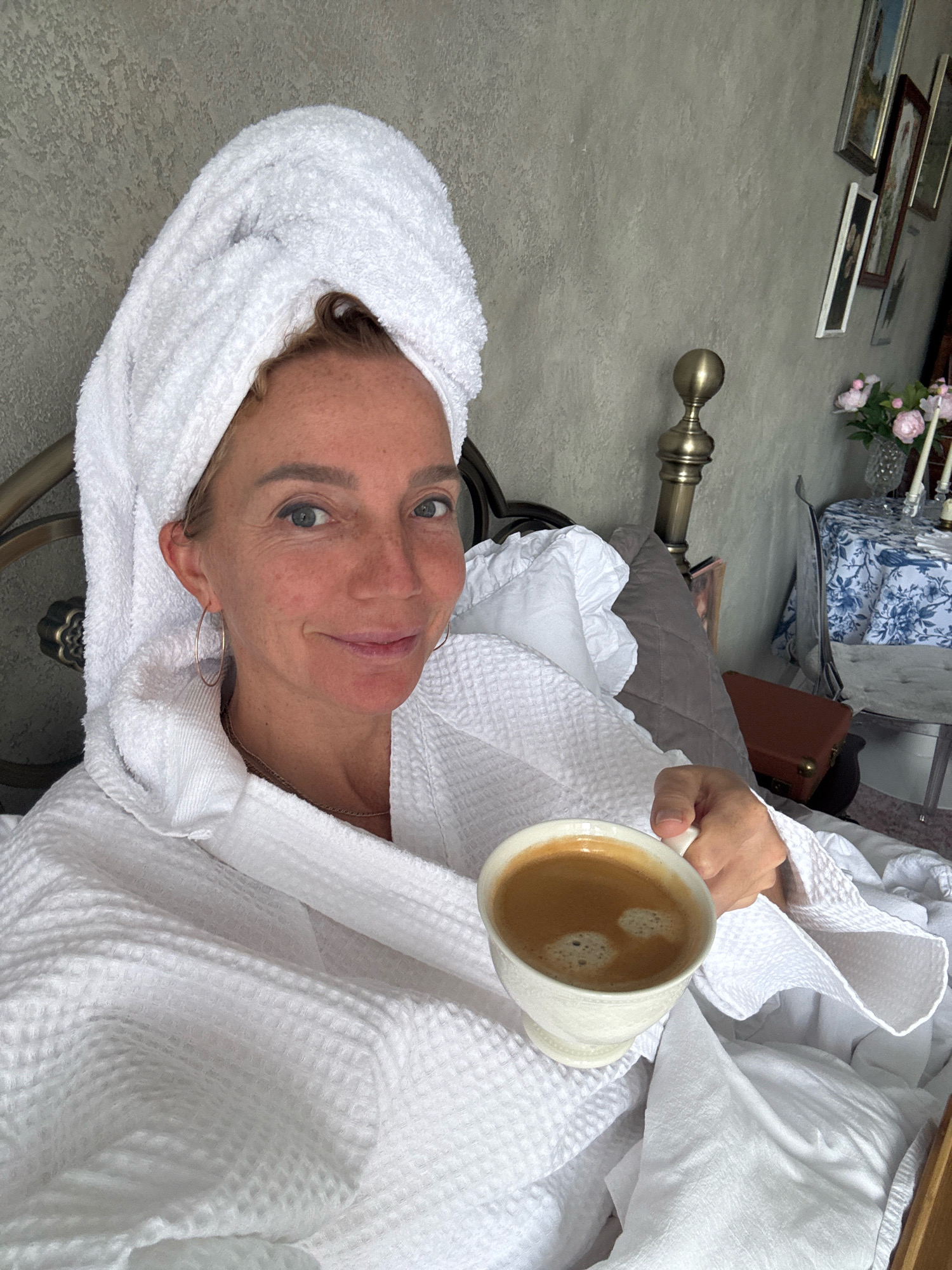 A woman is holding a cup of coffee while sitting in bed, wearing a white bathrobe and a towel wrapped around her head. The room appears to be a bedroom, with a bed, a bedside table, and framed pictures on the wall. The overall impression is one of relaxation and comfort. The location is not specified in the image.