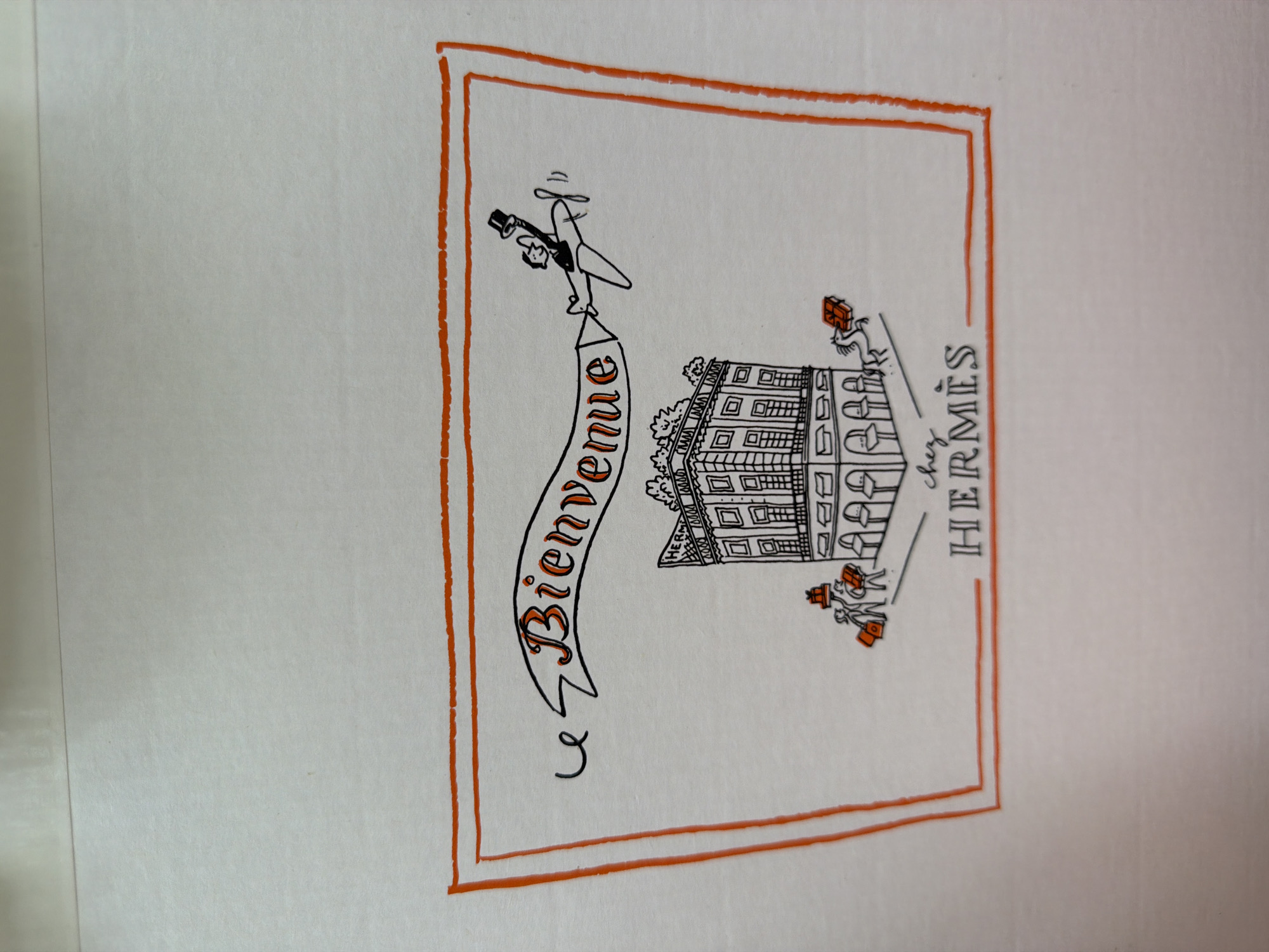 A drawing on white paper depicts a festive scene within an orange border. A banner across the top reads "Bienvenue," with a figure in a top hat piloting a small airplane to the right of the banner. Below, a whimsical building is rendered with detailed windows and an ornate facade. Two small figures carrying shopping bags stand to the left of the building, while a horse with a gift box balances on its back to the right. Below the building, the text "chez HERMÈS" is written. The drawing style is characteristic of a postcard or greeting card, with simple lines and a charming, illustrative quality. There is no discernible information within the image that suggests a location in Penzano, Italy.