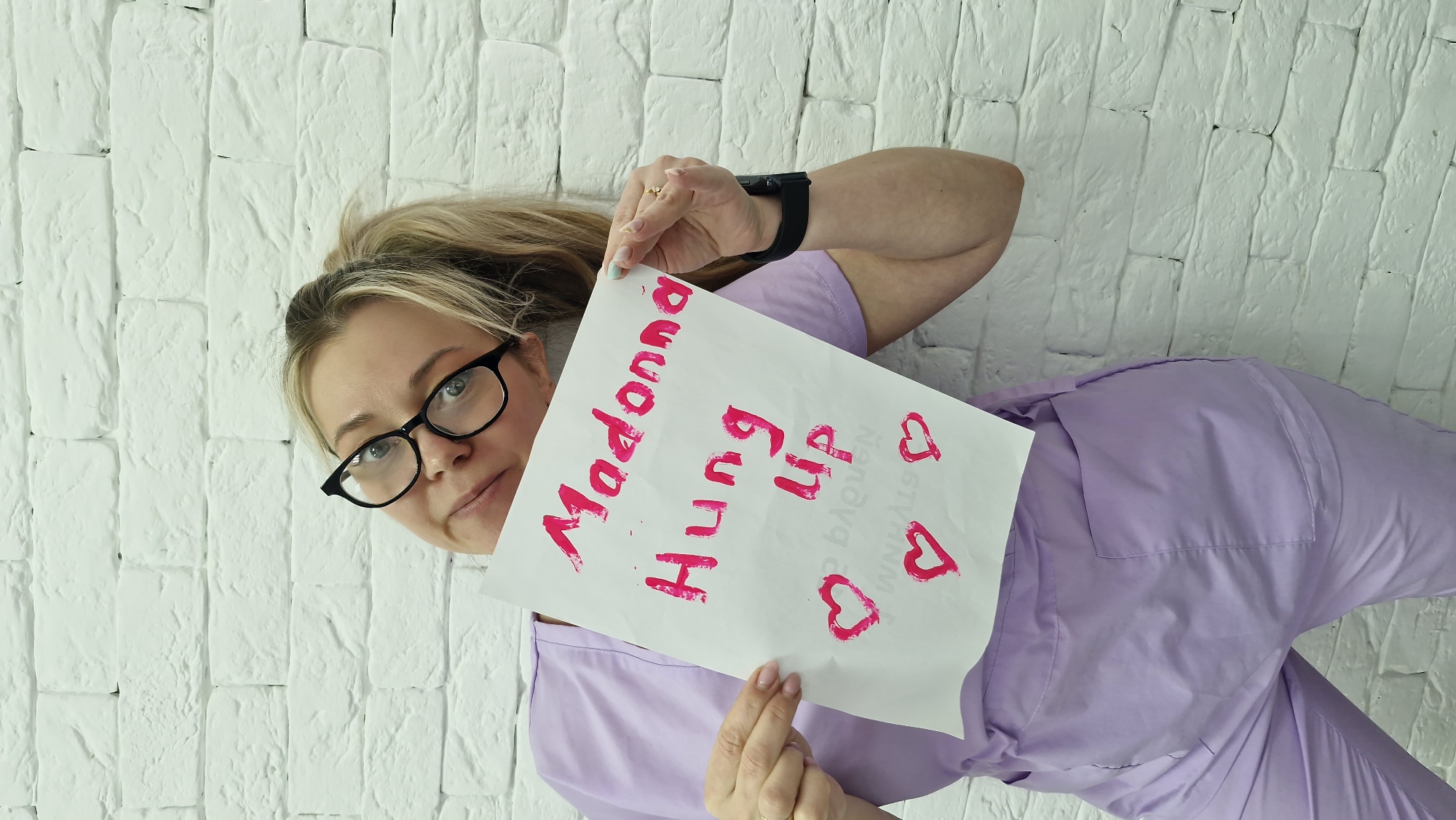 The image, rotated 90 degrees counter-clockwise, depicts a blonde woman with black-rimmed glasses, dressed in a light purple uniform or scrubs. She is positioned against a textured white brick wall, looking directly at the viewer with a neutral to slightly curious expression, peering over her glasses.
She holds a white sheet of paper with prominent pink handwritten text. Her left hand, adorned with a black smartwatch, grips the top of the paper, while her right hand, wearing a ring, supports the bottom. Her fingernails are manicured, some painted a light blue, others a light pink or natural shade.
The text on the paper clearly reads:
"Madonna
Hung
Up"
Below this, two pink heart symbols are drawn. Fainter, blurred text is also visible further down and to the right, which appears to include "ANABAYOU" and "BAGUN" in a different style.
The setting is an indoor space, indicated by the white brick wall, likely a studio or office environment. The location context is Otchor, Russia.