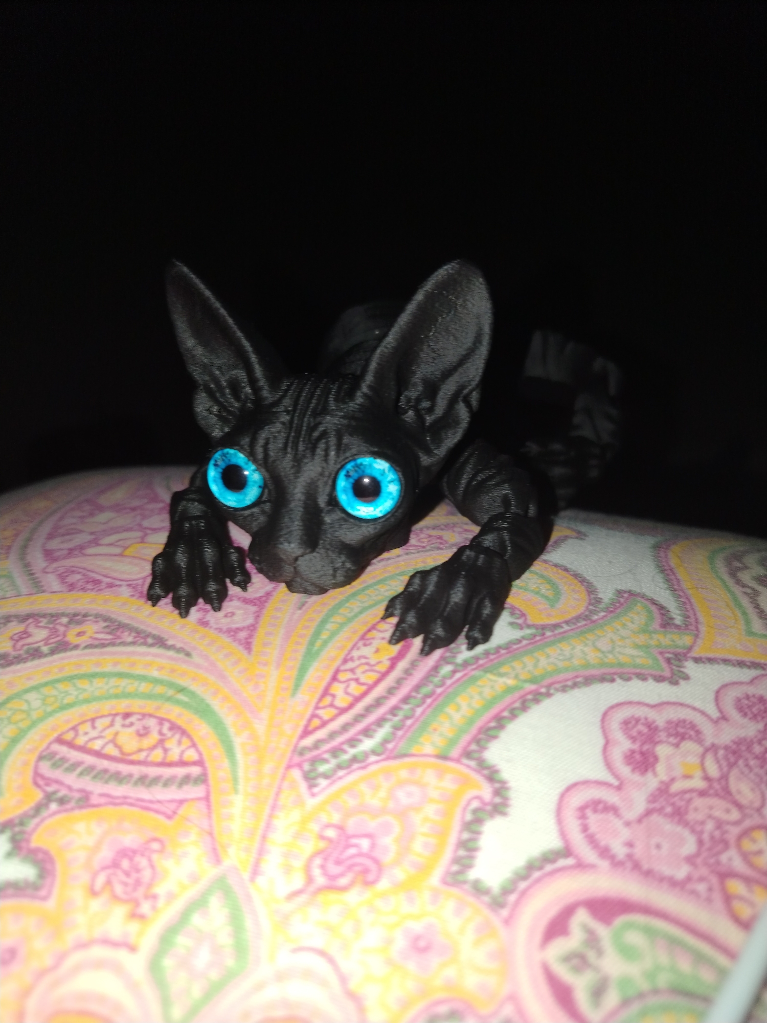 A black, Sphynx-like cat figurine with bright blue eyes lies on a pillow or cushion with a paisley pattern featuring pink, yellow, and green hues. The figurine has visible striations, suggesting it might be 3D printed. The background is dark. There is no indication of this image being taken in Izhevsk, Russia.