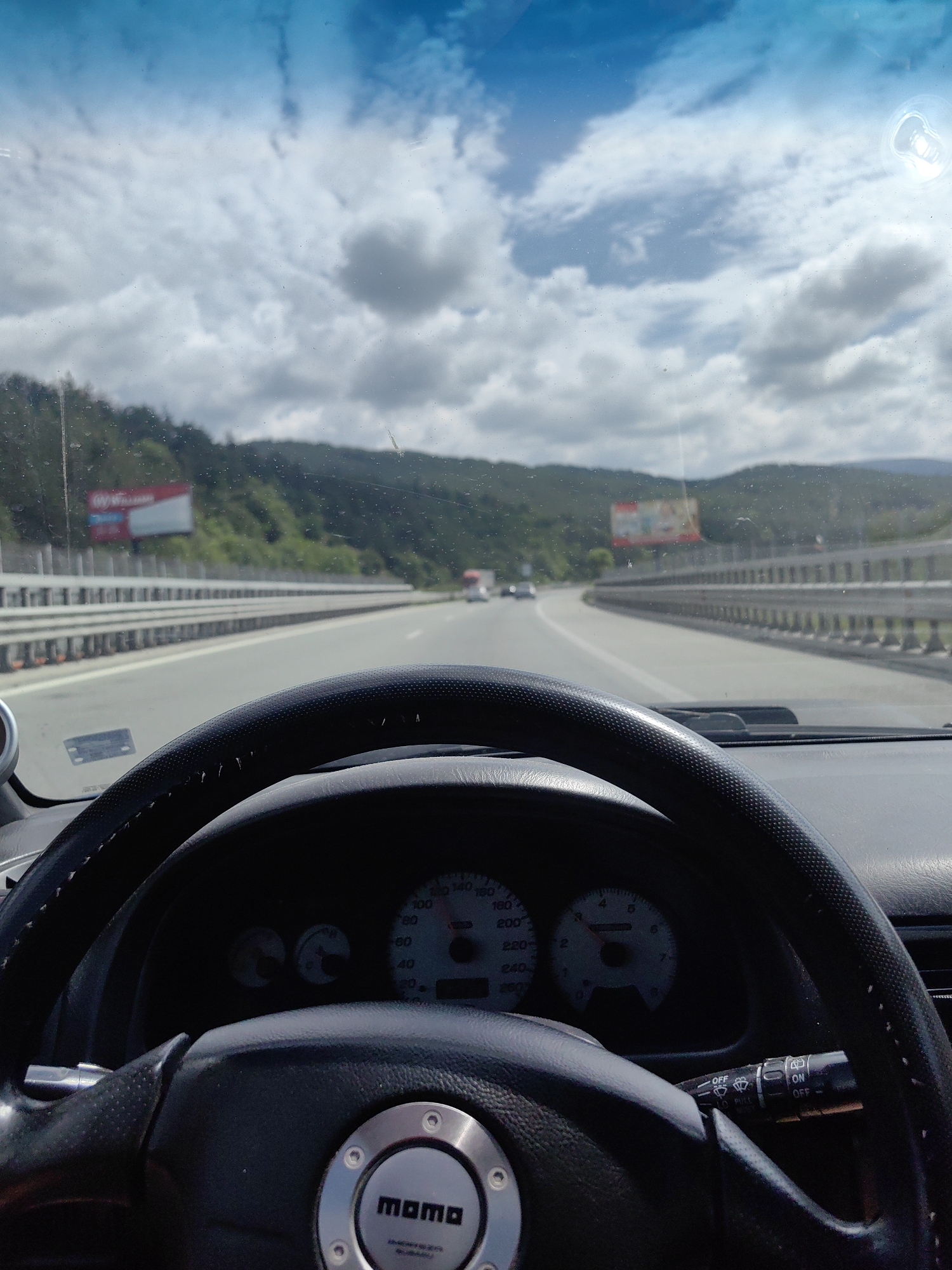 The image shows a first-person perspective from inside a car, driving on a highway. The steering wheel, dashboard, and speedometer are visible in the foreground. The speedometer indicates a speed between 80 and 100 km/h. The road ahead curves to the right, with guardrails on either side. In the background, there are hills covered with trees and some billboards. The sky is partly cloudy, with bright sunshine. The scene appears to be a typical highway drive, possibly in a rural or semi-rural area. The provided location, Potop, Bulgaria, is a small village, and the image likely depicts a road in or near that location.
