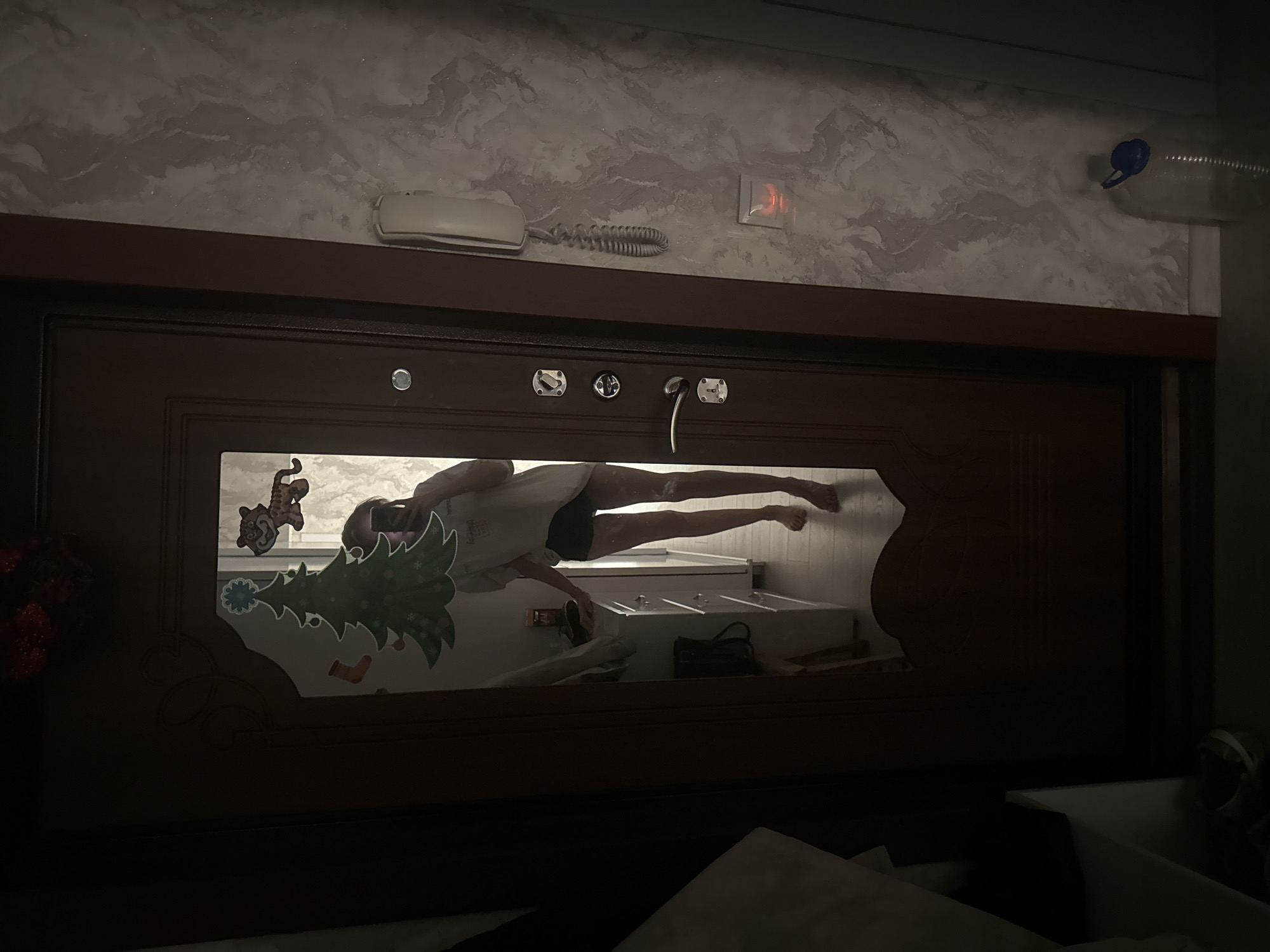 The image shows a bedroom with a large dark wooden headboard in the foreground. The headboard has a rectangular opening with a reflective surface, possibly a mirror, which shows a bathroom scene. Inside the bathroom, there's a person wearing a white shirt and dark shorts, appearing to be leaning over a white sink area, potentially washing something. Decorations like a Christmas tree sticker and a cartoon tiger sticker are visible on the mirror surface.
Above the headboard, the wall is textured with marble-like patterns. A white telephone handset, a coiled cord, a light switch, and a plastic water bottle can be seen attached to the wall. The overall lighting is dim, with the brighter area being the reflection in the headboard's mirror.
The city of Izhevsk, Russia, is not discernable from the image.