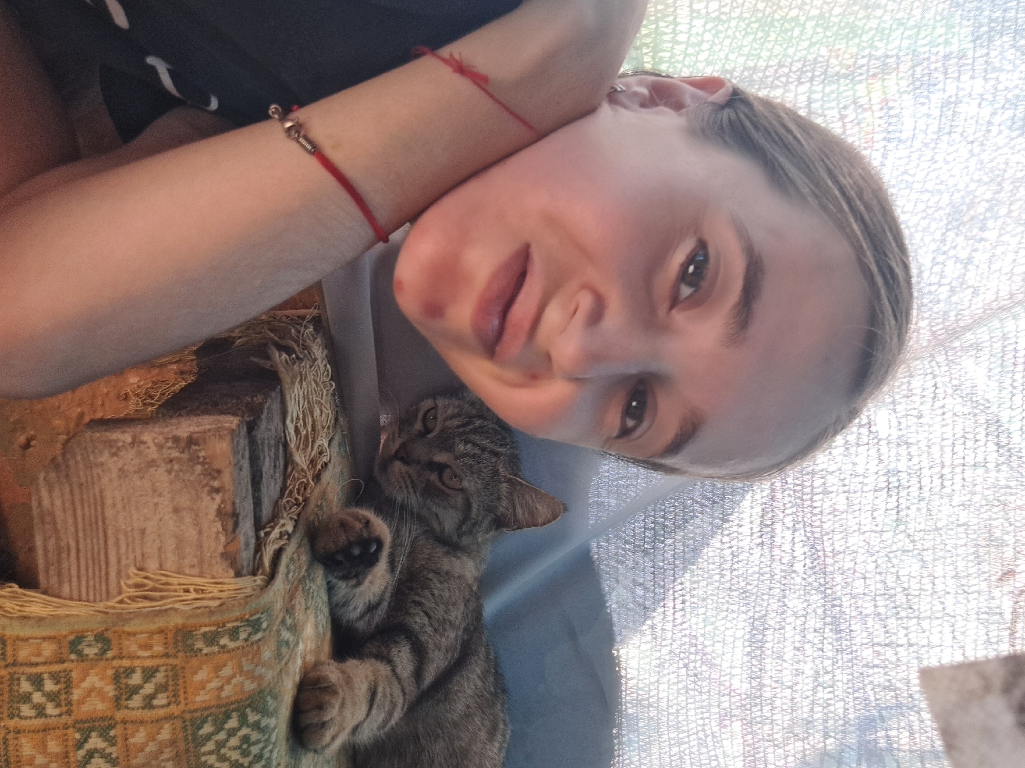 In Vypasne, Ukraine, a close-up selfie shows a person with light brown hair pulled back, a small hoop earring in their left ear, and wearing a black t-shirt. Red bracelets are visible on their left wrist. Their right arm is around their neck. To the right of the person, a tabby cat is lying down on a wooden surface covered with a patterned fabric that has yellow and green elements and fringe. The background is obscured by a light-colored, textured netting. The lighting suggests an outdoor setting during the day.