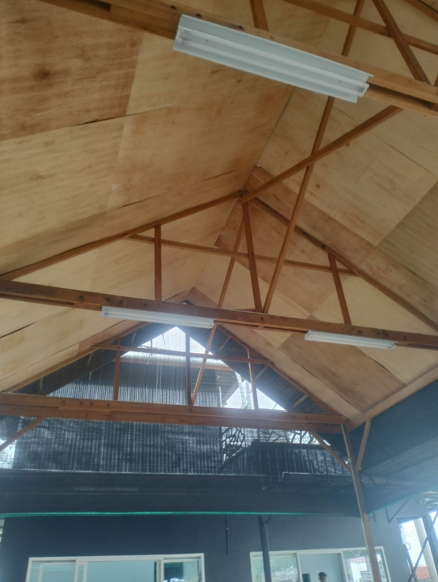 The media file shows the interior of a structure with a wooden roof made of plywood panels. A triangular wooden framework supports the roof, with several fluorescent light fixtures attached to the beams. The structure appears to be an open-air or semi-open space, as indicated by the presence of corrugated metal and black mesh material on the sides, which might be for ventilation or shade. Below the roof, a dark-colored wall is visible, with white-framed windows on the lower level. The lighting suggests it could be daytime, with natural light filtering in from the sides. The overall scene depicts a utilitarian space, possibly a workshop, market stall, or covered outdoor area, with a focus on the construction and lighting elements of the ceiling. There is no visible text.