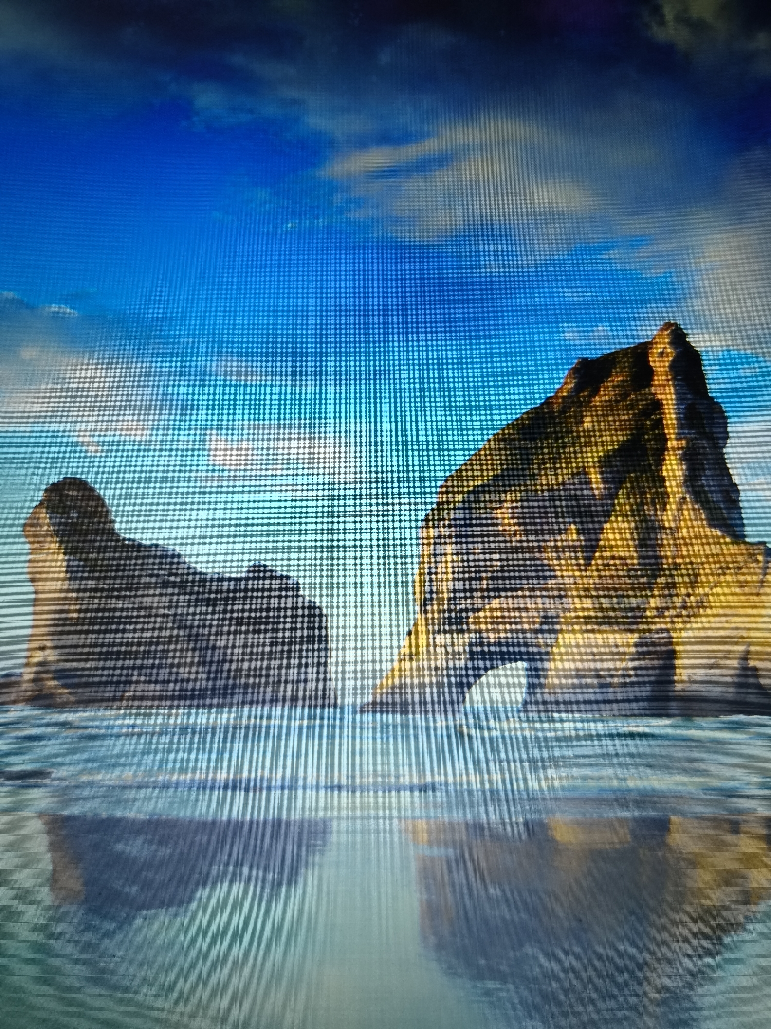 The image depicts a coastal landscape with two prominent rock formations rising from the water. The rock formation on the right has a natural archway carved through it. The sea reflects the rocks and the sky. The sky is blue with scattered white clouds. There is no evidence to place this scene in Kryvyi Rih, Ukraine, or any city at all. It is a seaside location. There are no visible people, objects, or actions apart from the natural environment.