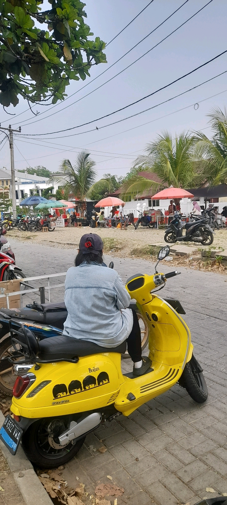A rear view shows a person seated on a yellow scooter in Jonggol, Indonesia. The scooter is adorned with a "The Beatles" logo and a silhouette of the band members. The person is wearing a light blue denim jacket, dark pants, and dark shoes with white socks. The scooter has a license plate with the text "B 6522 VWA" and "06-28". In the background, there is a building with a sign that reads "SEKOLAH AMAL KASIH". Several other motorcycles are parked in the area, and various stalls with colorful umbrellas are set up, suggesting a market or gathering. Palm trees and foliage are visible, indicating a tropical environment. The ground is paved with interlocking bricks and dirt. The sky is overcast.