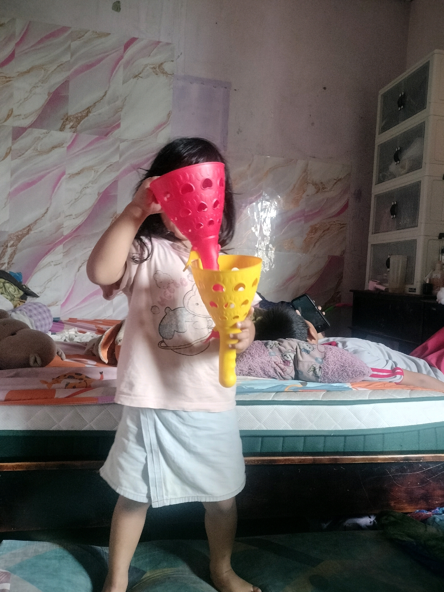 A young child, possibly a girl, stands in the foreground of a bedroom, holding two brightly colored plastic cups, one red and one yellow, over her face. She is wearing a light-colored t-shirt with graphics and a light blue wrap skirt. To the right, another child is lying on a bed, looking at a smartphone. The bedroom appears to be in a residential setting, with a patterned wallpaper on the back wall and a stack of white storage drawers to the right. The room is cluttered with various items, including stuffed animals and bedding. The lighting suggests it is daytime. The exact location within South Jakarta, Indonesia, cannot be determined from the image alone.
