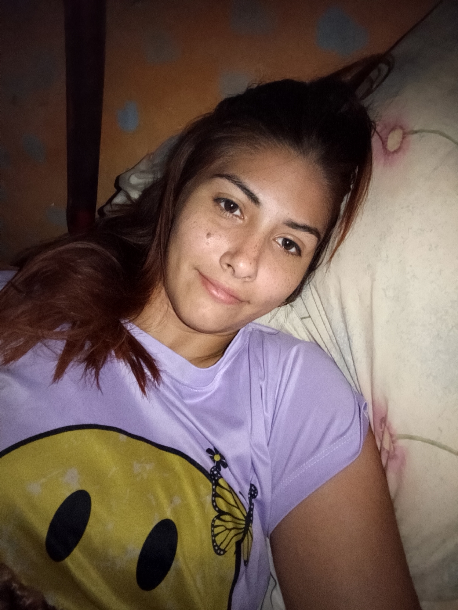 A young woman lies on a pillow. She wears a lavender T-shirt with a large, yellow smiley face and butterfly design. Her brown hair is partially pulled back. Her complexion is fair, with noticeable freckles. The background suggests an interior setting, possibly a bedroom. The location, Juan Griego, Venezuela, cannot be determined from the image alone.