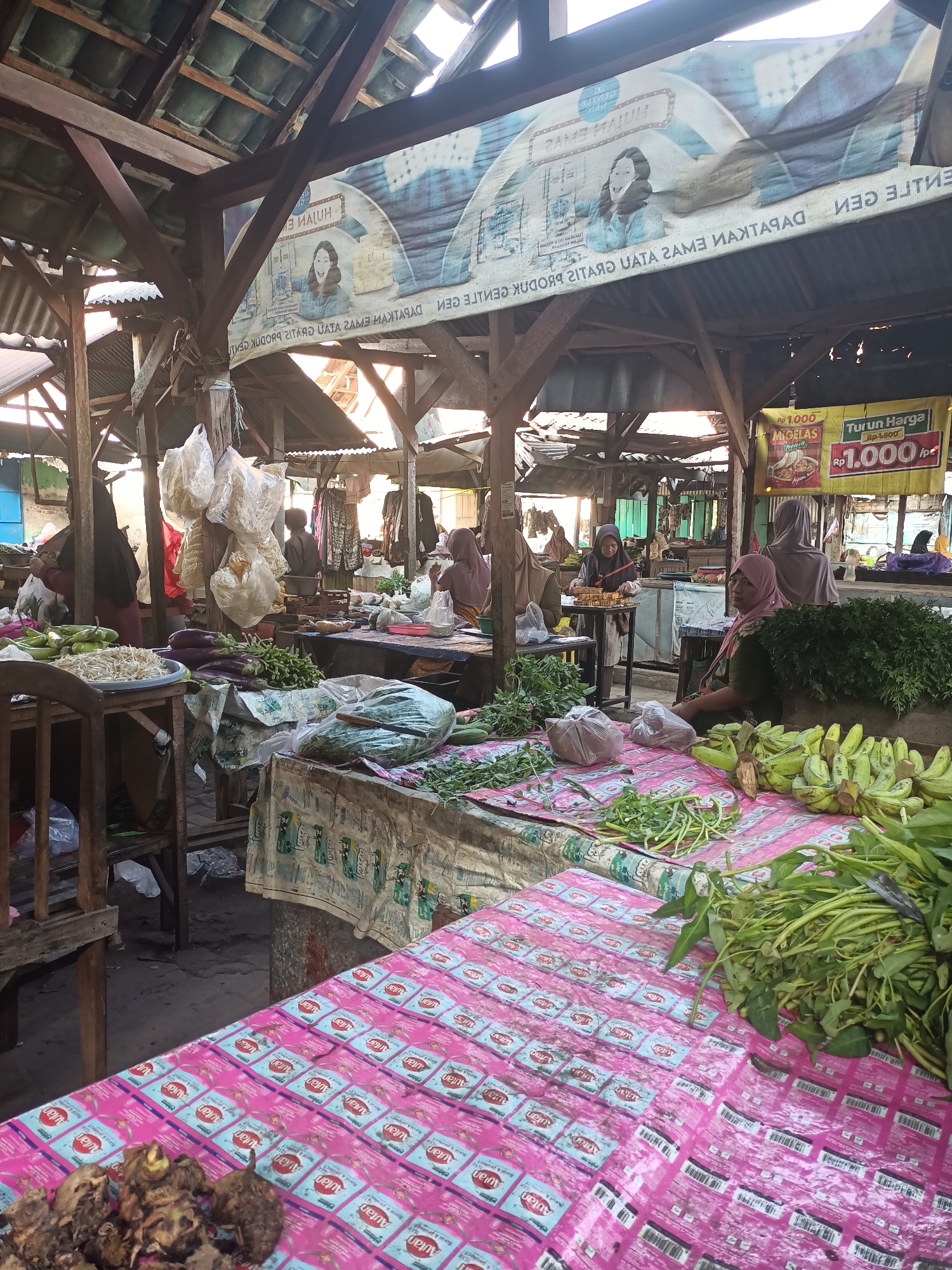 This image depicts a bustling outdoor market scene in Badas, Indonesia. The market is covered by a weathered wooden roof supported by beams, with a corrugated metal roof visible in some sections. The foreground is dominated by various fresh produce displayed on tables covered with colorful cloths. Bunches of bananas, leafy greens, and what appear to be taro root are prominent. In the background, several individuals are seen engaged in market activities. Some are attending to stalls, while others appear to be customers. Clothing styles suggest a Muslim cultural context, with many women wearing hijabs. The overall atmosphere is one of a vibrant local marketplace, likely with a focus on agricultural goods.