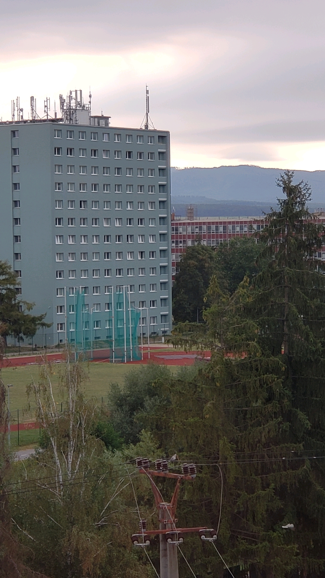 The image shows a view of a residential building with many windows, a sports field with a green net, and trees, with a cloudy sky and hills in the background. This appears to be a view of a dormitory building at the University of Prešov in Prešov, Slovakia. The building is tall and rectangular, with a light blue facade and rows of white-framed windows. On the roof, there are several telecommunications antennas and equipment. In front of the building, there is a sports field with a running track and a green net structure, possibly for a sport like volleyball or tennis. Several trees, including a large evergreen tree, are visible in the foreground and middle ground. In the distance, a range of hills can be seen under a hazy sky. A utility pole with electrical insulators is also visible in the foreground. There are no people visible in the image.