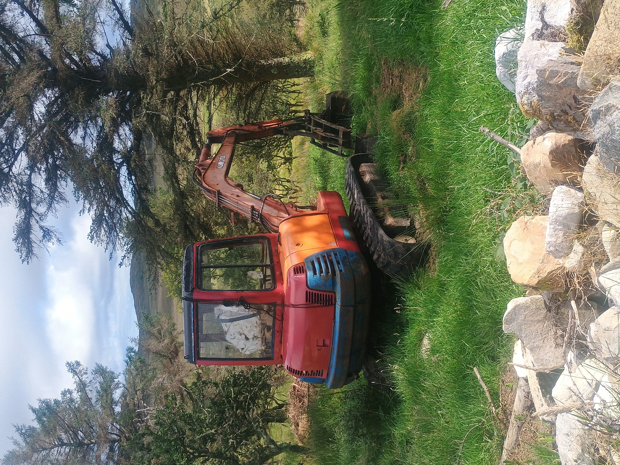 An orange and red Kubota excavator sits idle in a grassy, uneven field. The excavator has a red cab with a white bag inside and a blue and black front grill. Its arm is extended with the bucket lowered. The scene is surrounded by trees, some with bare branches, and in the background, there are rolling hills under a cloudy sky. In the foreground, a pile of large, light-colored rocks and some dried vegetation is visible. The ground is covered in green grass.
