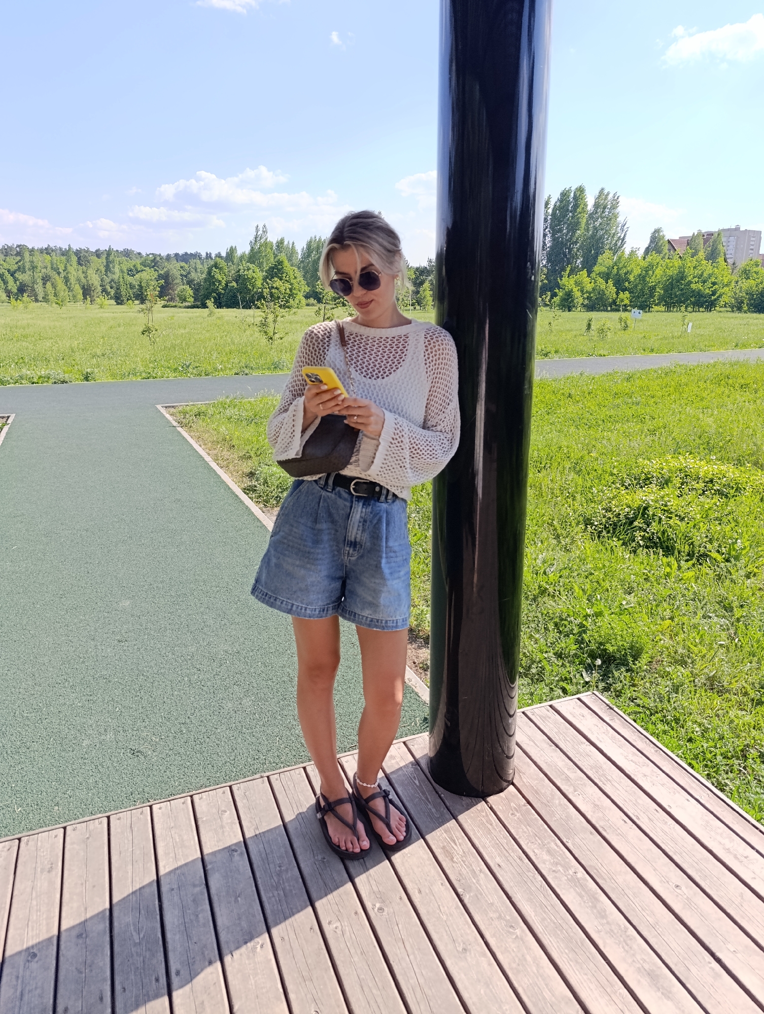 A woman stands in a park in Unknown, Unknown, holding a yellow smartphone. She wears sunglasses, a white crochet sweater, high-waisted denim shorts with a black belt, and black sandals. A small, dark purse hangs from her shoulder. She leans against a tall, shiny black pole positioned on a wooden platform. Beyond the platform is a green, paved path leading to a grassy area dotted with trees. In the distance, a building is visible on the right. The sky is blue with scattered clouds.