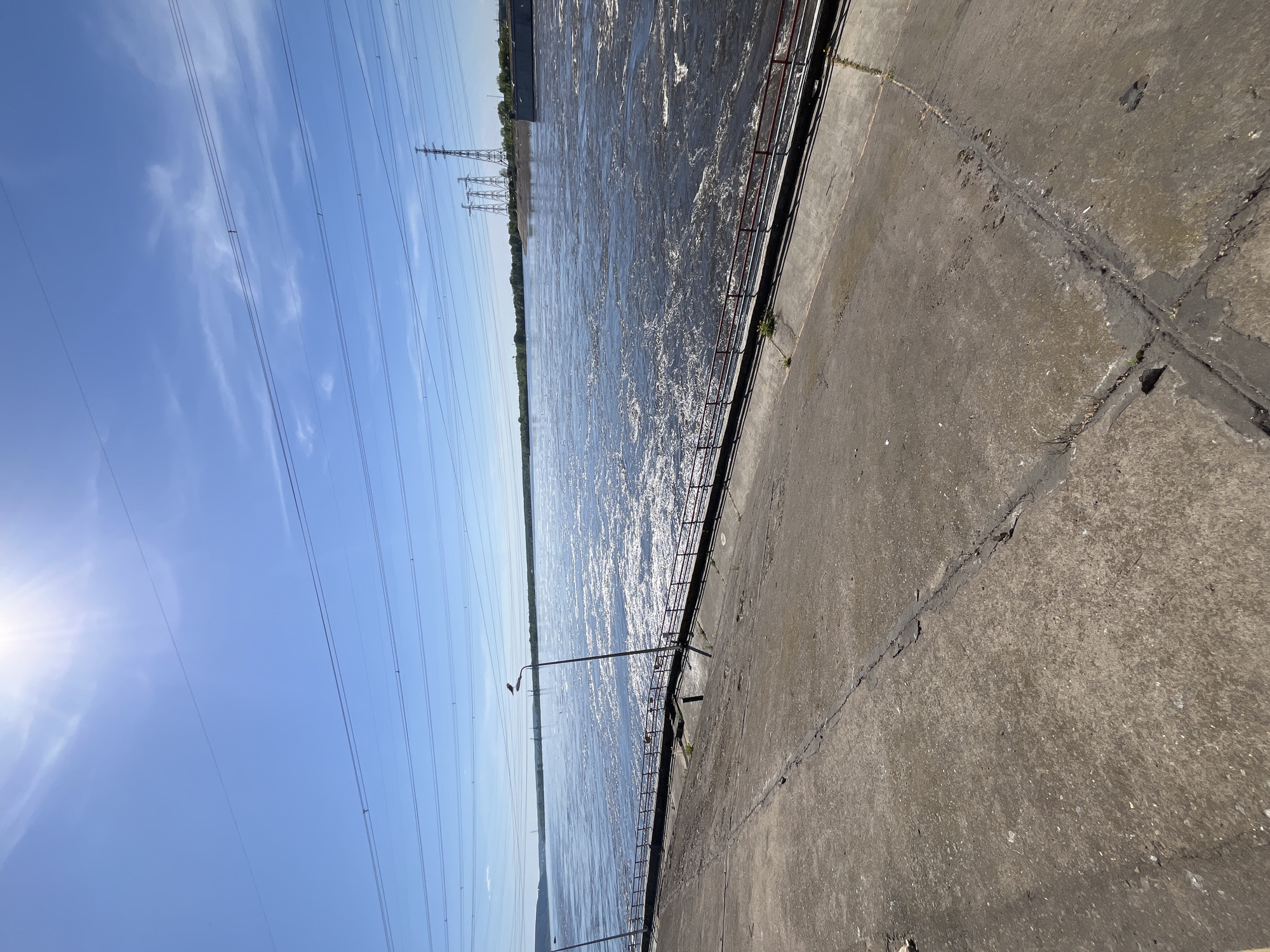 The image depicts a concrete embankment adjacent to a wide body of water, likely a river or reservoir, under a bright blue sky with some clouds. Overhead power lines stretch across the sky. A metal railing runs along the edge of the embankment. In the distance, power pylons are visible on the opposite bank. The concrete surface of the embankment has cracks and patching. The image orientation is rotated counter-clockwise. Given the city specification, this is likely the Volga River in Noviy, Russia, although this cannot be confirmed with the provided information. There are no people visible in the image.