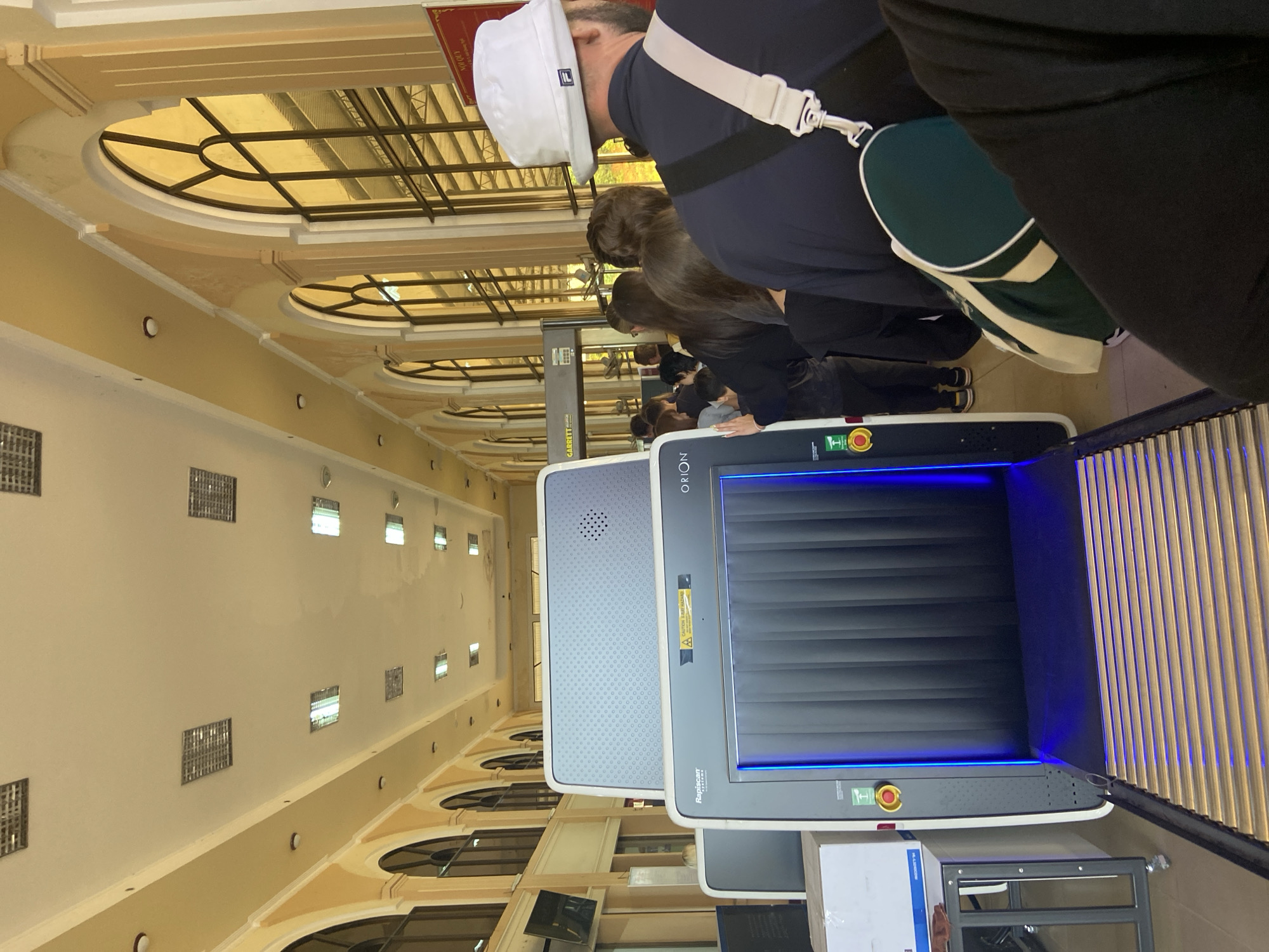 This is a full-frame, eye-level, medium shot of a security checkpoint at a transportation hub in Bo Y, Vietnam. The scene depicts a queue of people waiting to pass through a baggage scanner and a metal detector. In the foreground, a Rapiscan Orion baggage scanner is visible with its blue interior lighting. A conveyor belt extends from the scanner. To the right, a man wearing a white bucket hat and a dark shirt is standing in line, with a green and white duffel bag on his shoulder. Behind him, a line of diverse individuals, varying in age and gender, are patiently waiting. Further down the line, a Garrett metal detector archway is present. The environment appears to be an indoor terminal with arched windows and yellow-toned walls, characteristic of colonial architecture. The lighting is bright and evenly distributed.