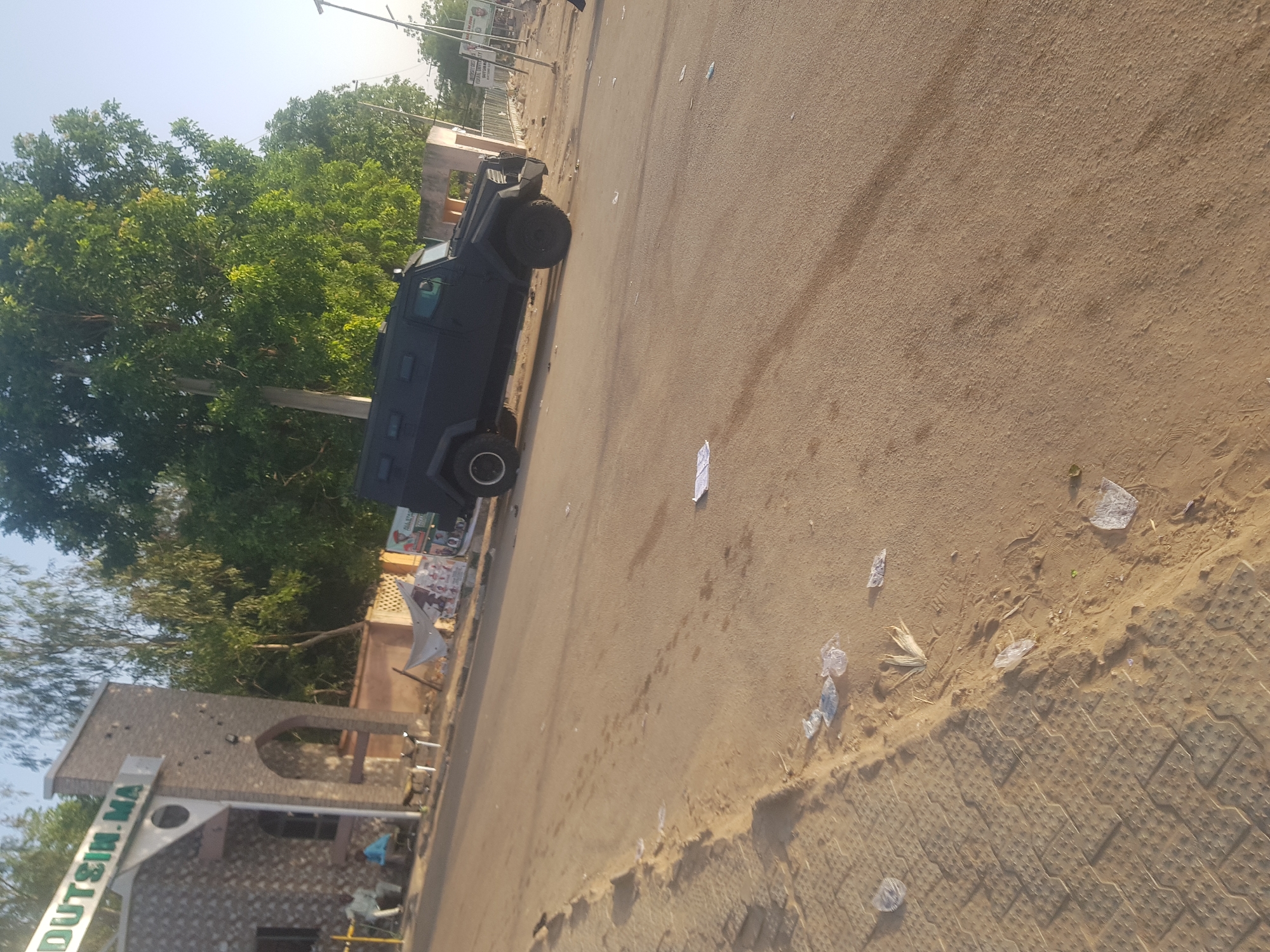 A dark-colored armored vehicle is parked on a paved road in what appears to be a semi-urban environment. The road is lined with trash. A stone building with a sign indicating the location as "DUTSIN-MA" is on the left side of the frame. In the background, there are trees and other buildings. The environment suggests a daytime scene in Dabawa, Nigeria.