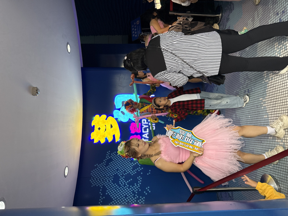This image depicts an indoor scene within an entertainment or attraction venue located in Shinan District, China. The environment features a curved blue wall adorned with illuminated yellow and blue Chinese characters, notably "梦幻" (Dream/Fantasy), and the English text "TACYP" beneath them, alongside a dotted world map graphic. The ceiling is white, equipped with recessed lighting fixtures and a smoke detector. The floor is covered with light blue tiles arranged in a grid pattern.
In the foreground to the left, a young adult female entertainer stands, dressed in a sleeveless pink top with feathered trim and a voluminous, layered pink tulle skirt, paired with white socks and platform sneakers. Her hair is styled in braids, topped with a colorful floral headpiece. She holds a white and blue sign displaying Chinese characters that translate to "Qingdao, a romantic city, plus love."
To her right, a young girl, wearing a red and black plaid shirt over a white top, light blue wide-leg jeans with butterfly embroidery, and silver sneakers, stands facing slightly right. Behind this child, a person in a vibrant clown costume, featuring a plaid top, striped pants, and a multi-colored afro wig, holds yellow and orange balloon animals.
On the right side of the frame, an adult female is seen from behind, wearing a black and white vertically striped shirt with a black yoke and black pants. She carries a black shoulder bag with a chain strap and observes the scene. In the bottom-left corner, a child's hand in a yellow sleeve is partially visible, reaching up from behind a red barrier rope. The overall scene suggests a family-friendly event with costumed performers interacting with visitors.