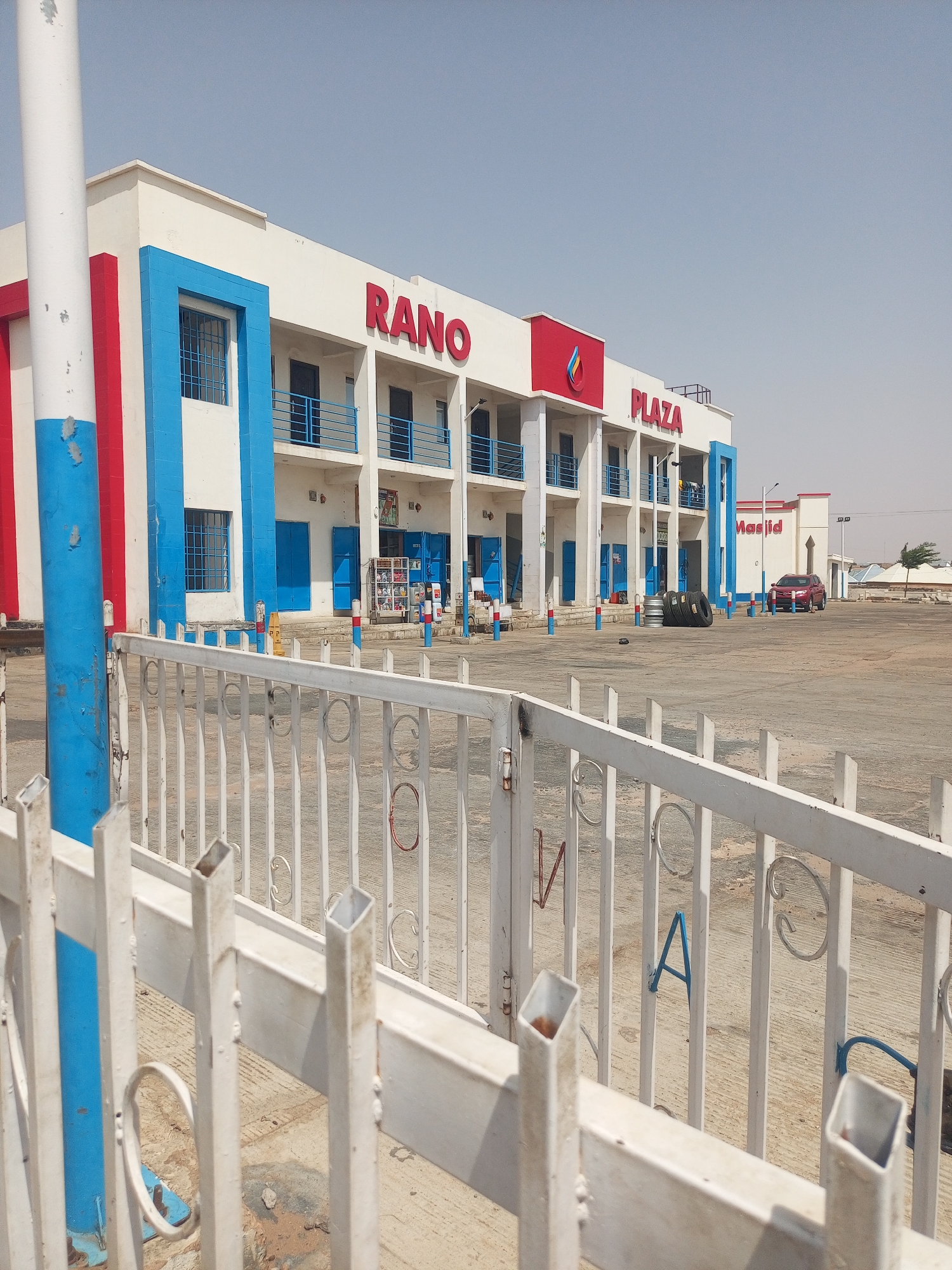 The image depicts a daytime view of Rano Plaza in Gombe, Nigeria. A two-story white building with blue and red accents houses multiple storefronts. The words "RANO PLAZA" are displayed in red lettering on the building's facade. A blue and red striped pole stands prominently to the left of the building. A large, paved parking area lies in front of the building. A white metal fence with circular decorative elements is visible in the foreground, partially obscuring the view. A red car is parked near a building with "Masjid" on it in the background.