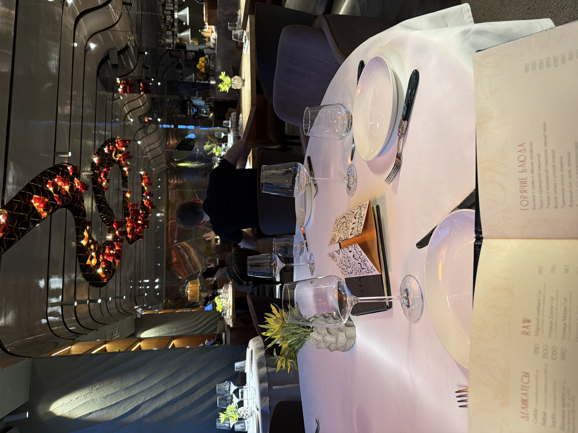 This media file captures the elegant interior of a modern restaurant in Moscow, Russia, likely during the evening due to the ambient lighting. The perspective is from a table set for dining, looking towards the back of the establishment.
In the foreground, a white tablecloth covers a round table, meticulously set with white plates, wine glasses, water glasses, and polished silverware including forks and knives. A small, spiky green plant in a textured gray pot serves as a centerpiece. A menu, open to pages displaying Cyrillic text, is prominently placed on the table, leaning against a dark cover.
The midground features other dining tables with chairs, some of which are occupied. A person in a black shirt, possibly a waiter or staff member, is visible from behind, attending to something near a table further back. The head of another person, presumably a diner, is partially visible in the bottom left corner.
The background reveals the sophisticated ambiance of the restaurant. A striking decorative lighting fixture, resembling a winding, snake-like structure adorned with numerous small red lights, hangs from the ceiling. Modern wall panels with integrated lighting create a sleek aesthetic. Further in the distance, a bar area is visible, with bottles, glasses, and possibly lemons on display. The overall decor suggests a high-end dining experience.
Visible text on the menu includes Cyrillic headings such as "ДЕЛИКАТЕСЫ" (Delicacies), "RAW", and "ГОРЯЧИЕ БЛЮДА" (Hot Dishes). Below "ГОРЯЧИЕ БЛЮДА," some dish descriptions are partially legible, including "Каннеллони с угрем и черным трюфелем" (Cannelloni with eel and black truffle), "Паста с мраморной говядиной" (Pasta with marbled beef), and "Лапша с крабовым мясом, черный перец" (Noodles with crab meat, black pepper). Various prices, such as 950, 720, 1000, 1950, 80, 90, 120, 150, and 180, are listed alongside menu items.