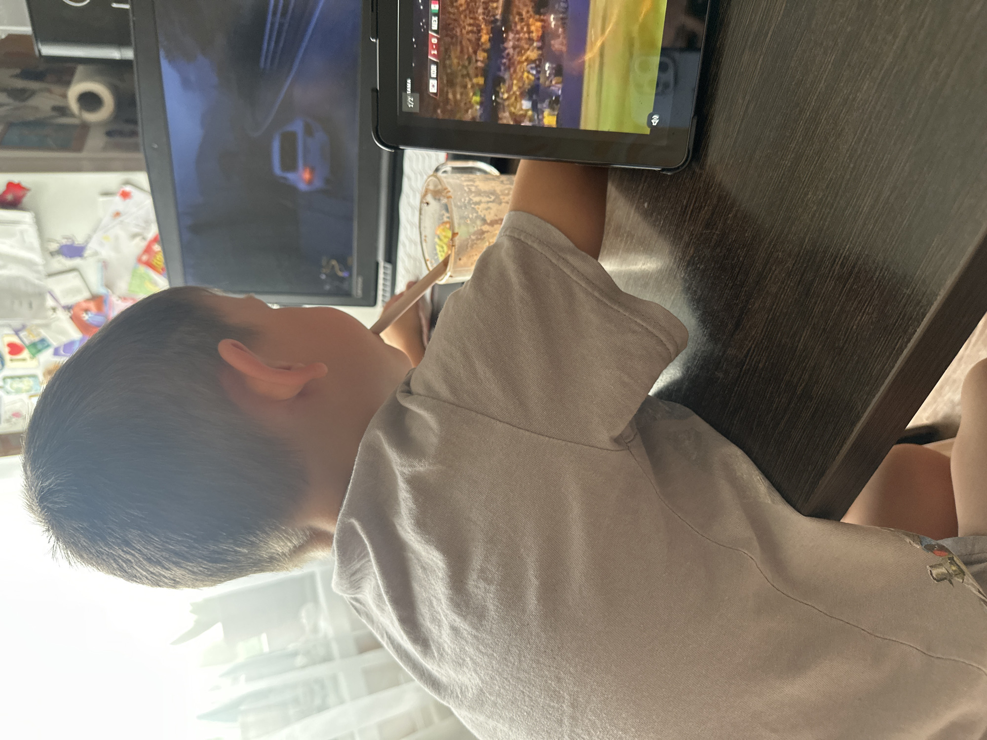 A young boy is sitting at a dark brown wooden table, facing away from the camera. He is wearing a light grey t-shirt and grey shorts. On the table in front of him, there is a laptop displaying a racing video game and a tablet showing a soccer match. A clear glass mug with a brown beverage and a straw is positioned between the boy and the laptop. The room appears to be brightly lit by natural light coming from a window on the left side of the frame, with a white curtain partially visible. The wall behind the boy is decorated with various colorful papers and drawings. The overall scene suggests a casual indoor activity, likely in a home environment.