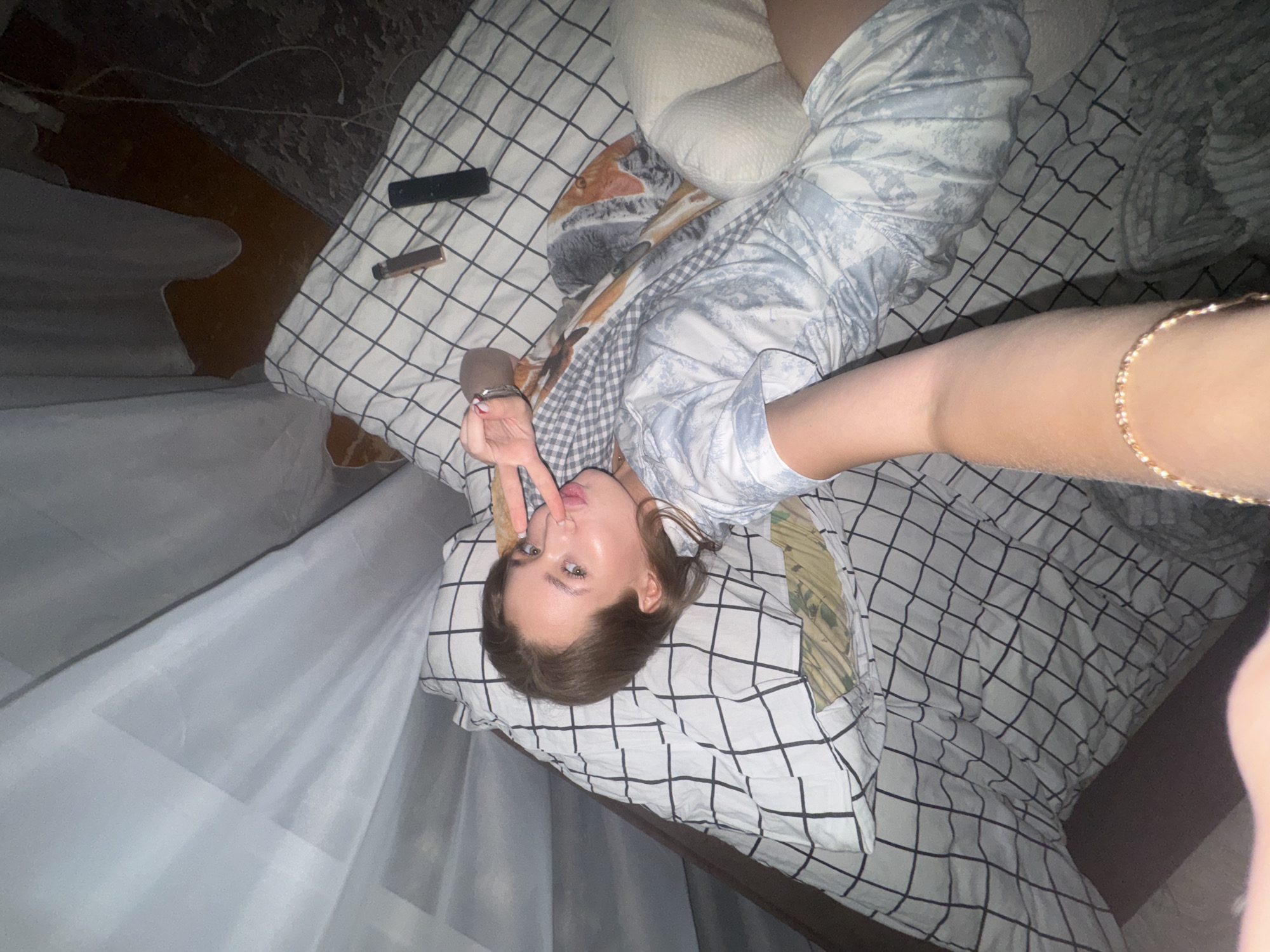 The media file shows a young woman lying on a bed with a black and white checkered comforter. She is looking at the camera with a playful expression, holding up two fingers in a peace sign. She is wearing pajamas with a light blue and white pattern. A pillow with a textured surface is behind her head. To the left of the bed, sheer white curtains are visible. On the bed near her are a remote control and a small, rectangular object that appears to be a lighter. The scene appears to be indoors, likely in a bedroom. The lighting suggests it could be nighttime or a dimly lit room. The overall mood is casual and relaxed. There is no visible text in the image. The location context is Chaikovsky, Russia.
