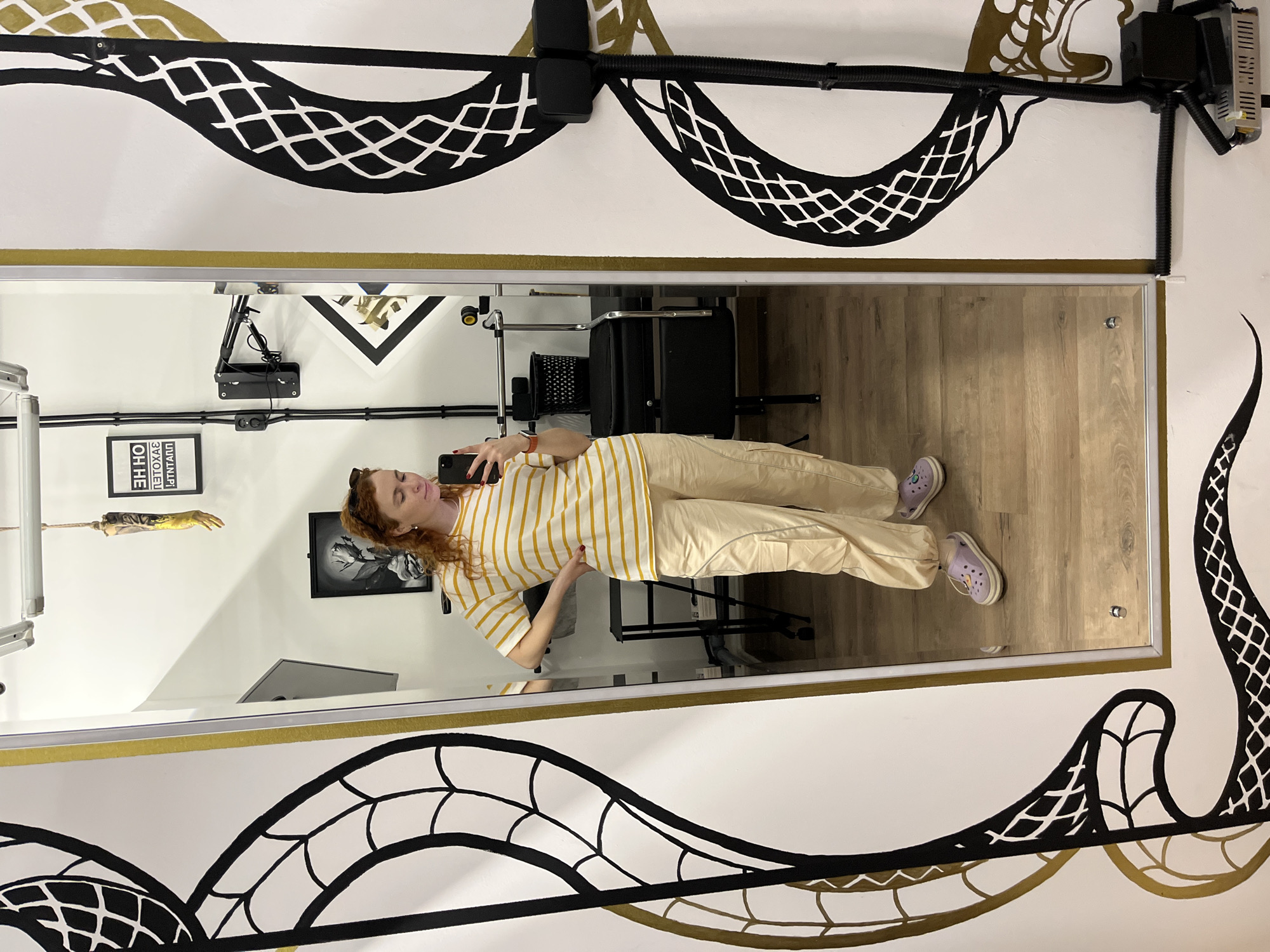 The image shows a woman taking a selfie in front of a mirror. She has red curly hair, is wearing a striped yellow and white shirt, tan pants, and purple shoes. The reflection shows she is in a room with wood flooring. Behind her in the reflection is a black chair and a small table. On the wall behind her, there are framed pictures and a model of a human arm hanging down. The wall outside of the mirror is decorated with a white background and a large black and gold wave pattern resembling a snake. The location is Chaikovsky, Russia.