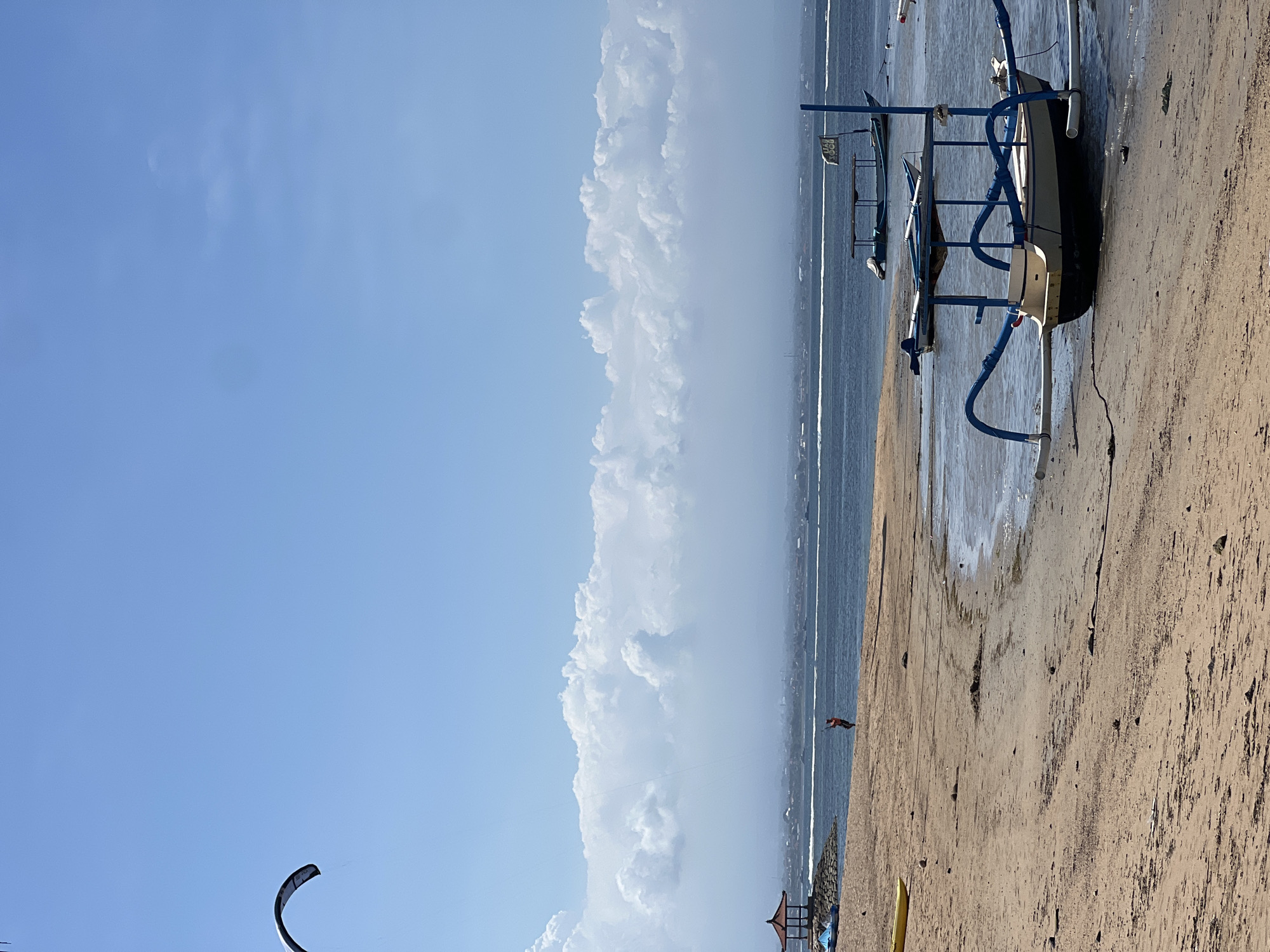 The image captures a serene beach scene in Denpasar, Indonesia. The foreground is dominated by a sandy beach with scattered debris. A traditional Balinese fishing boat, known as a jukung, rests partially on the shore, its distinctive outriggers adorned with blue accents. In the middle ground, the calm blue ocean stretches towards a distant coastline, dotted with several other boats. A person in an orange shirt is visible on the beach, engaged in kitesurfing, with a kite visible in the upper left corner of the sky. The sky is a clear blue, with a band of fluffy white clouds stretching across the horizon. A small, red-roofed pavilion is situated on a rocky outcrop on the left side of the image. The overall atmosphere is tranquil and evokes a sense of a typical day at the beach.