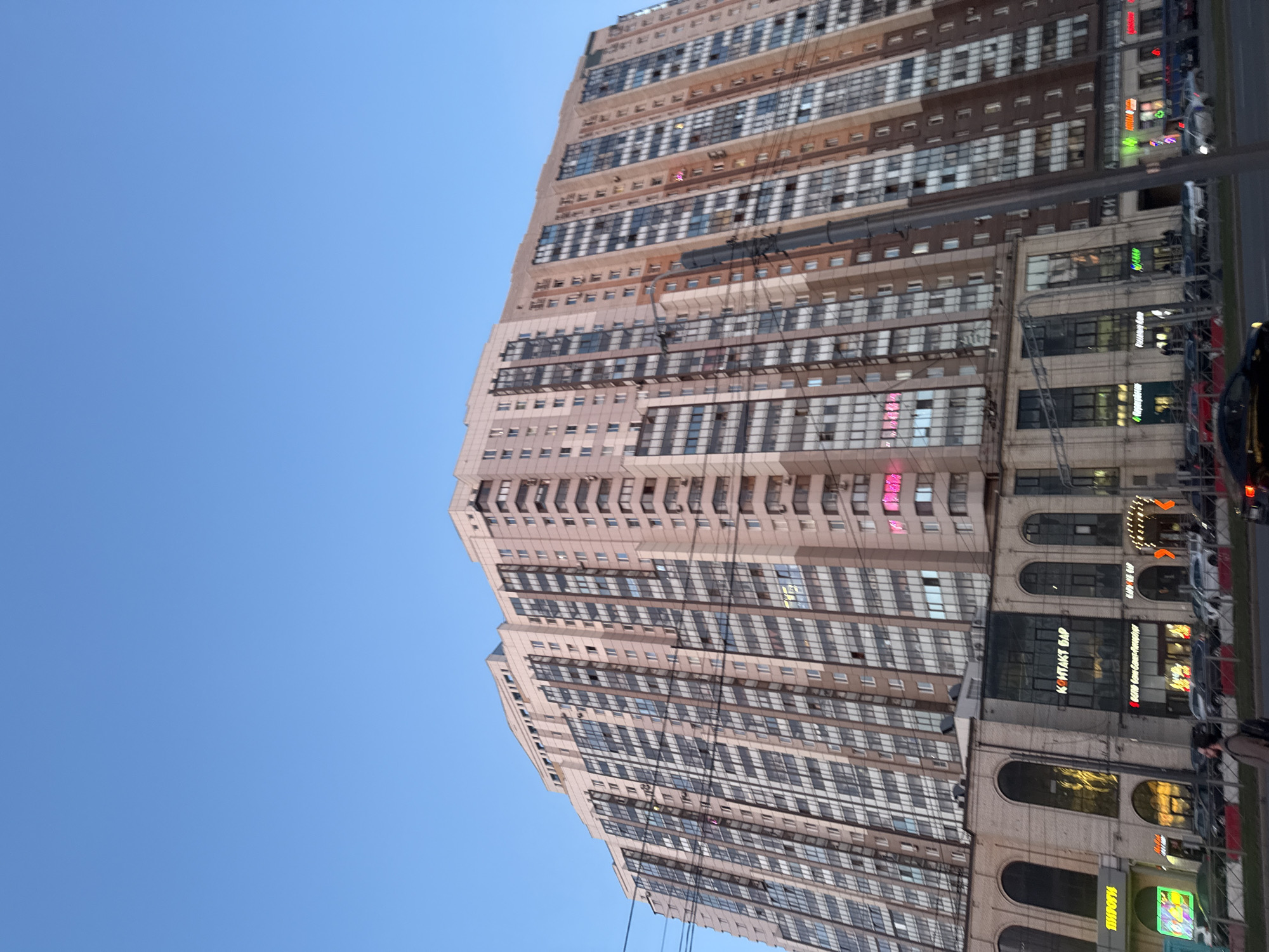 A tall, modern multi-story building dominates the right side of the frame, rising against a clear, bright blue sky. The building appears to be made of light-colored panels, possibly light brown or beige, with numerous windows arranged in horizontal bands across its facade. Some sections of the building feature different architectural styles or materials, creating a layered appearance with darker horizontal elements. Visible power lines or cables crisscross in front of the building, adding an urban element.
At the street level, the building features several commercial establishments. Various illuminated signs, predominantly in Cyrillic script, suggest the presence of shops and services. Notable visible text includes: "КОНТАКТ БАР" (Kontakt Bar), "ПРОДУКТЫ" (Groceries), "ЛЕНА", "Пятерочка" (Pyaterochka, a supermarket chain), "ВКУСВИЛЛ" (VkusVill, another supermarket chain), and multiple "АПТЕКА" (Pharmacy) signs, indicated by text and a green cross symbol.
The scene depicts an everyday urban environment in Saint Petersburg, Russia, likely during daytime given the clear blue sky and bright ambient light, although some shop signs are already illuminated, possibly indicating late afternoon or early evening. The overall weather appears clear and sunny. Cars are partially visible on the street beside the building, and faint figures of people can be discerned on the sidewalk, indicating pedestrian activity. The image is oriented vertically, with the building extending upwards towards the top of the frame.