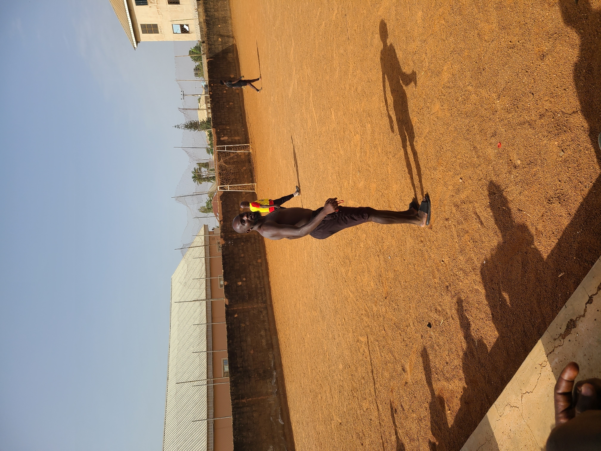 The scene shows a dirt field in Barakin Lerawi, Nigeria. A shirtless man in dark shorts and shoes is in the foreground. Another person is in the background. The shadow of a third person is also visible. A building and a fence are on the left side of the field. The sky is blue.
