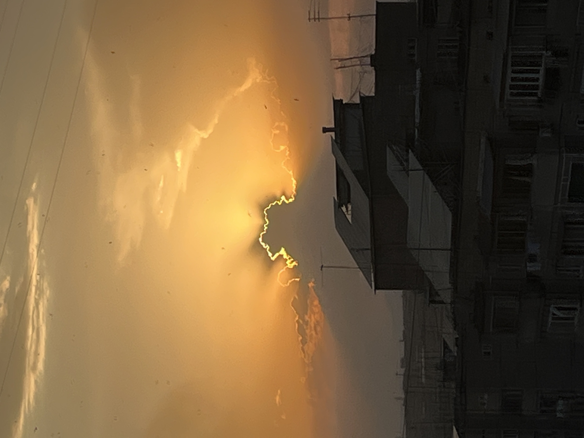 This is a photograph of a sunset over an urban environment, likely taken from a higher vantage point looking down. The sky is dominated by a vibrant orange and yellow hue, with rays of sunlight emanating from behind a thick, dramatic cloud formation. The cloud appears to be shaped in a way that resembles a silhouette. Below the sky, silhouetted buildings of varying heights fill the lower half of the frame. The architecture suggests a Soviet-era style, common in many parts of Yerevan, Armenia. The buildings are dark and indistinct due to the low light conditions. Some rooftops have various structures like vents, antennas, and possibly satellite dishes. The overall impression is one of a fading day in an urban landscape. There are no visible people or specific actions captured in the image. The scene is static and focused on the atmospheric effect of the sunset and the urban silhouette.