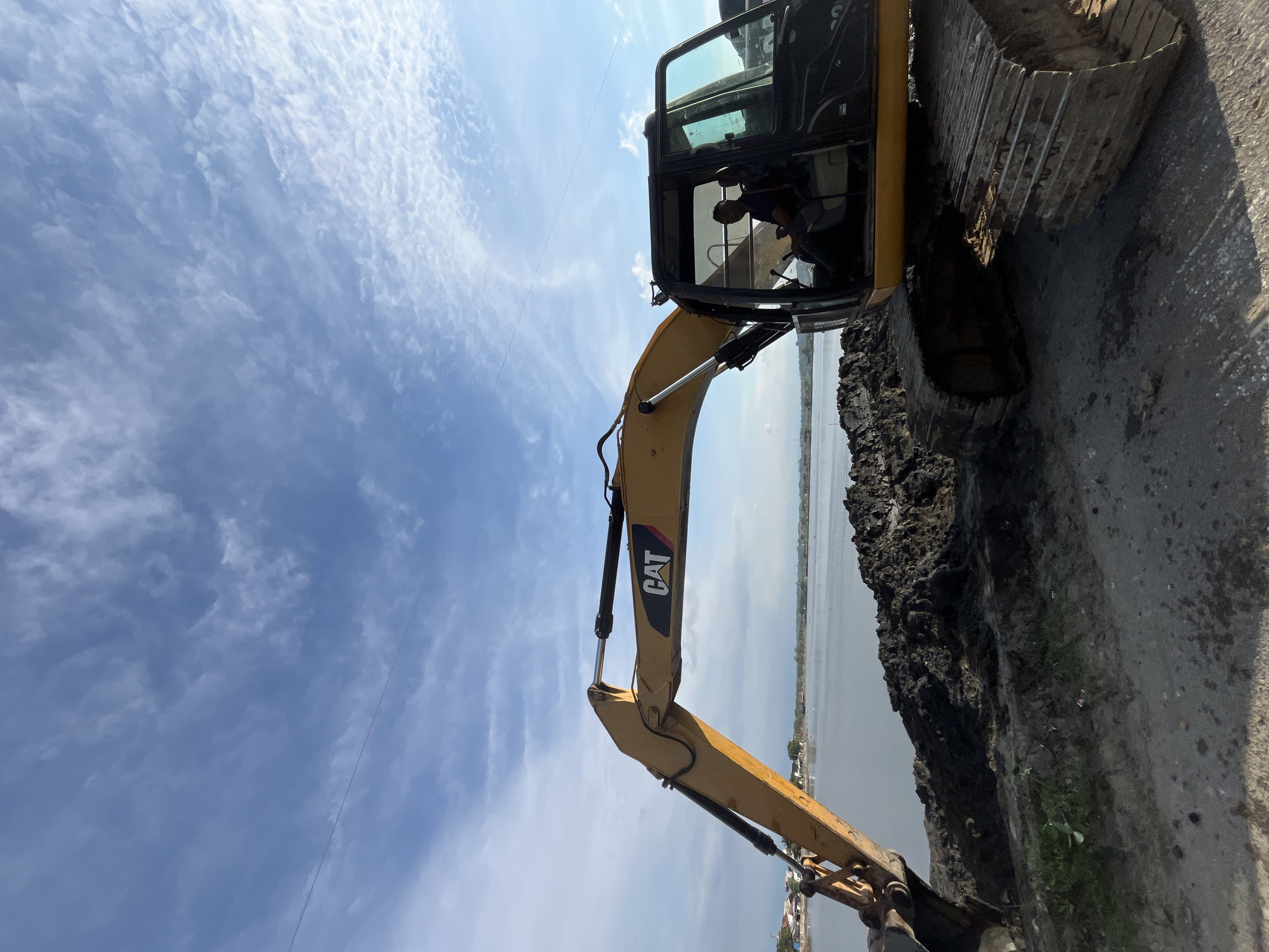 The image shows a yellow CAT excavator parked next to a pile of dark earth, likely mud or soil. The excavator is positioned on a paved surface. The boom extends towards a body of water in the background. The water appears relatively calm, and the opposite bank shows buildings, suggesting a coastal area or waterfront. The sky is blue with scattered clouds. Inside the excavator's cab, a person is visible, appearing to be the operator. Overhead power lines are visible against the sky. The location is Lhokseumawe, Indonesia.